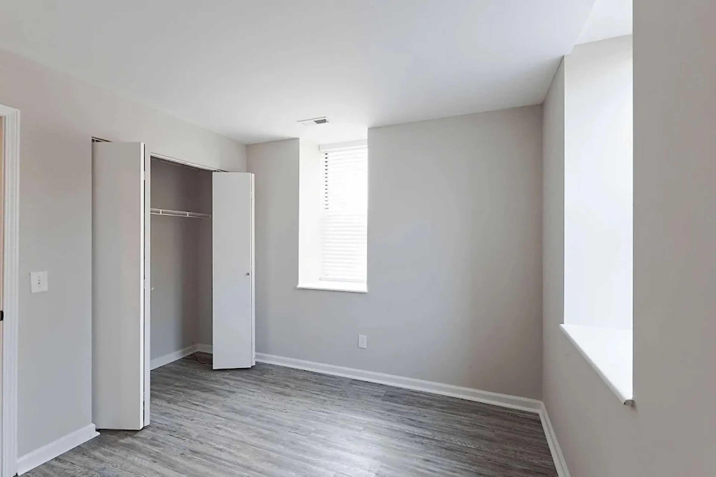 Empty room with light gray walls and a wooden floor. There is a small window with blinds and a doorless closet on one side. The space is bright, with natural light streaming in, creating a minimalist and modern atmosphere. No furniture or decorations are present, emphasizing the room's openness.