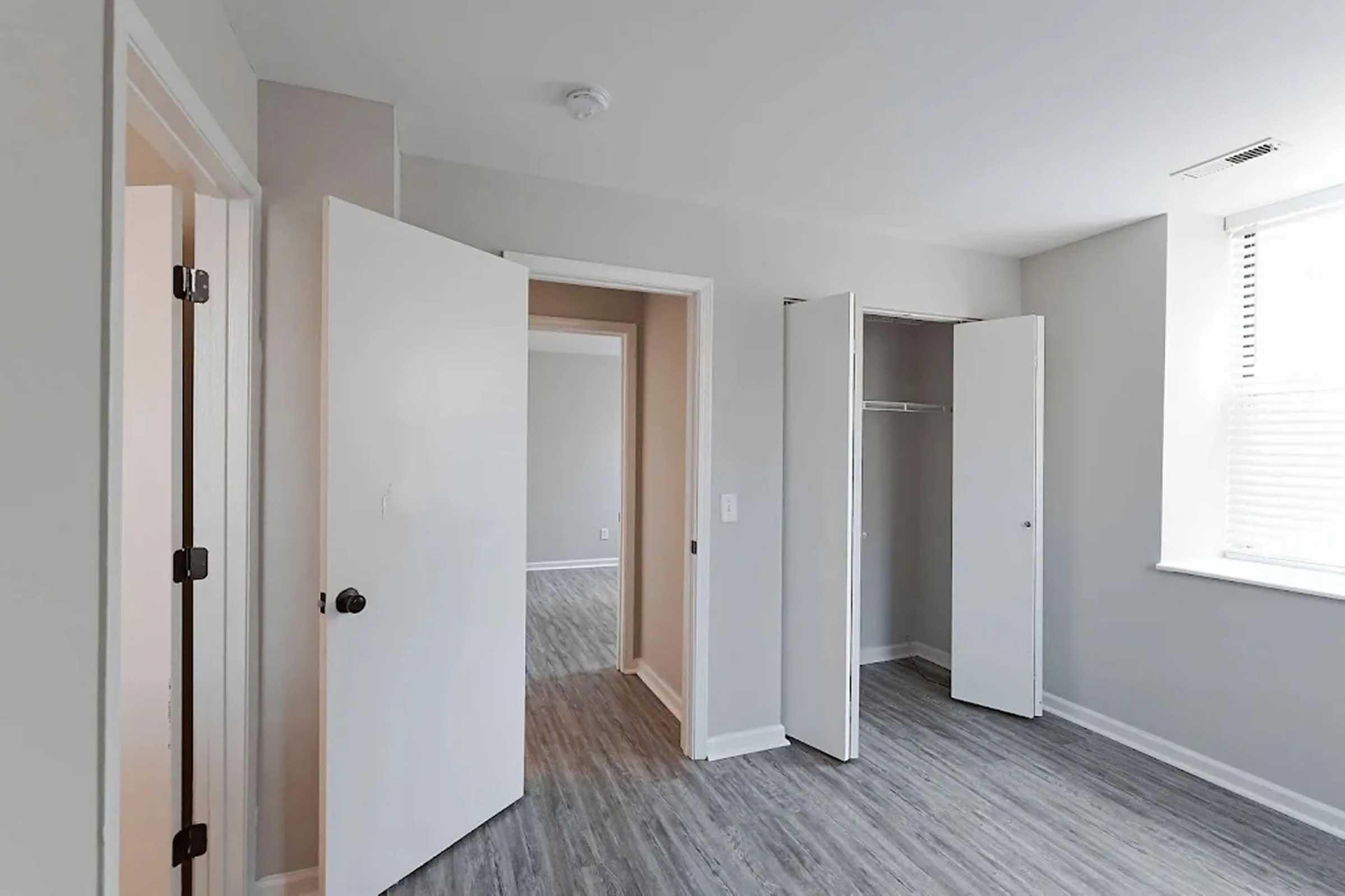 Interior of a room with light gray walls and vinyl flooring. There are two open white doors leading to another room and a closet, revealing an empty space with a window letting in natural light. The overall atmosphere is bright and minimalistic.