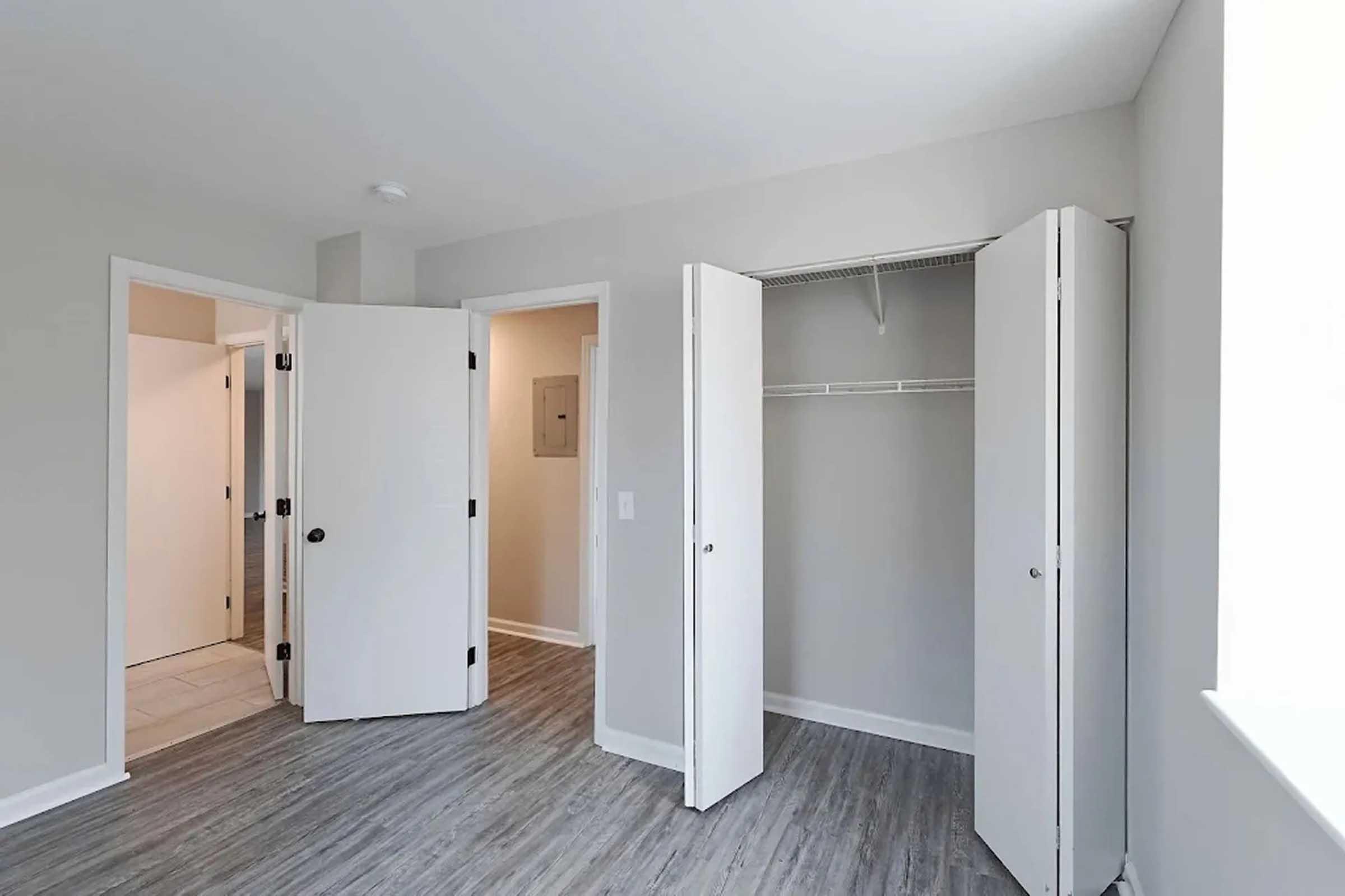 A bright and modern bedroom featuring light gray walls, hardwood flooring, and a closet with double doors. There is an entrance to a bathroom visible through one door, and ample natural light coming in from the window. The space is tidy and ready for decoration.