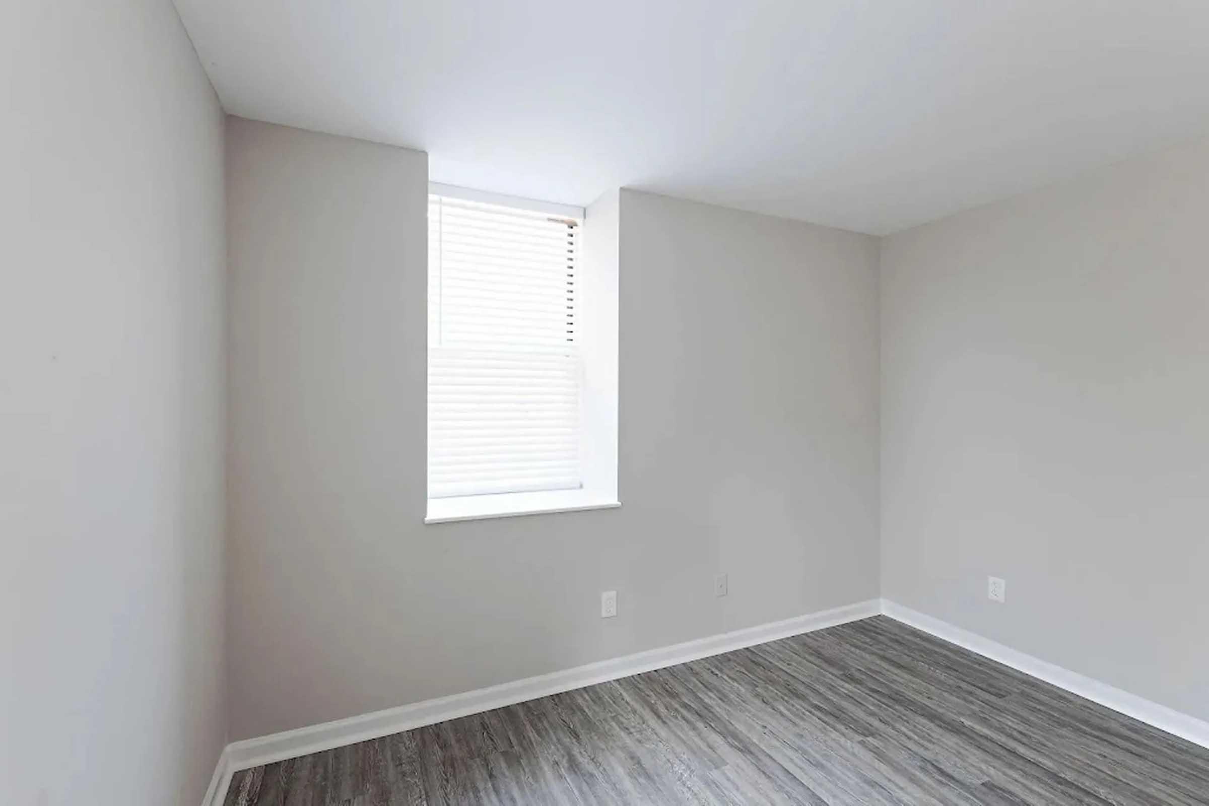Small, empty room with light gray walls and a window covered by blinds. The floor has a wood-like appearance, and there is natural light coming in from the window. The space is unadorned, creating a minimalist atmosphere.