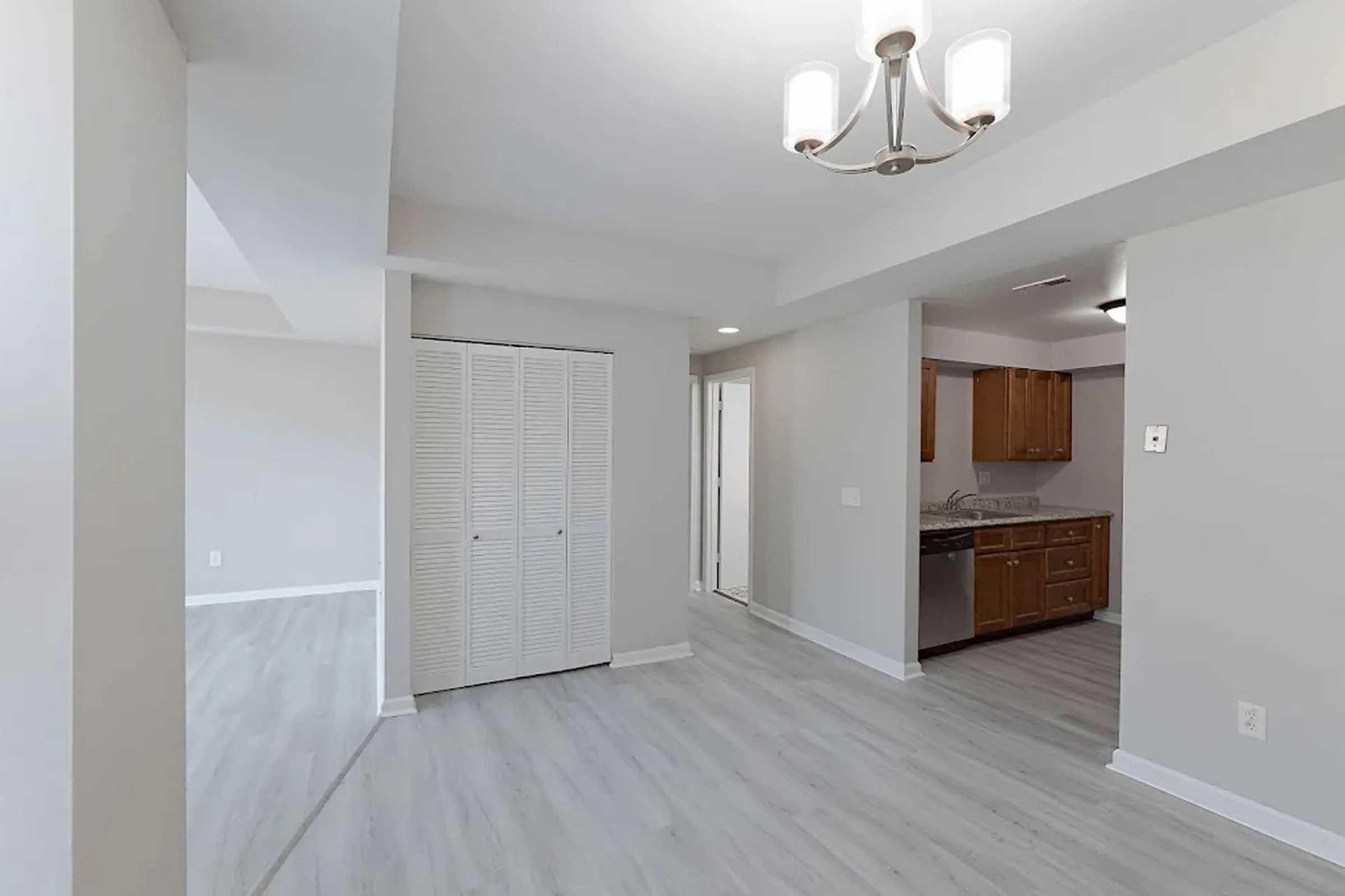 A light-filled room featuring a modern chandelier, gray walls, and light wood flooring. On the left, there's a white closet with sliding doors, while to the right is a doorway leading to a kitchen area with wooden cabinets and stainless steel appliances. The overall space is open and inviting.