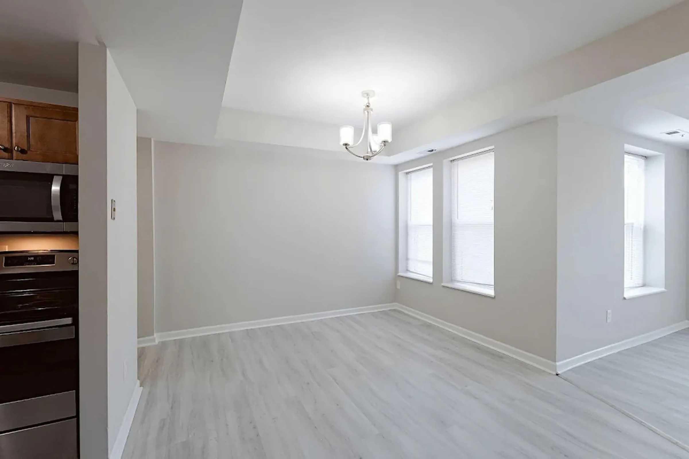 A well-lit, empty room featuring light gray walls, white trim, and a chandelier. There are two windows allowing natural light, and the flooring is a light-colored laminate. A kitchen area with modern appliances, including an oven and microwave, is visible on the left.