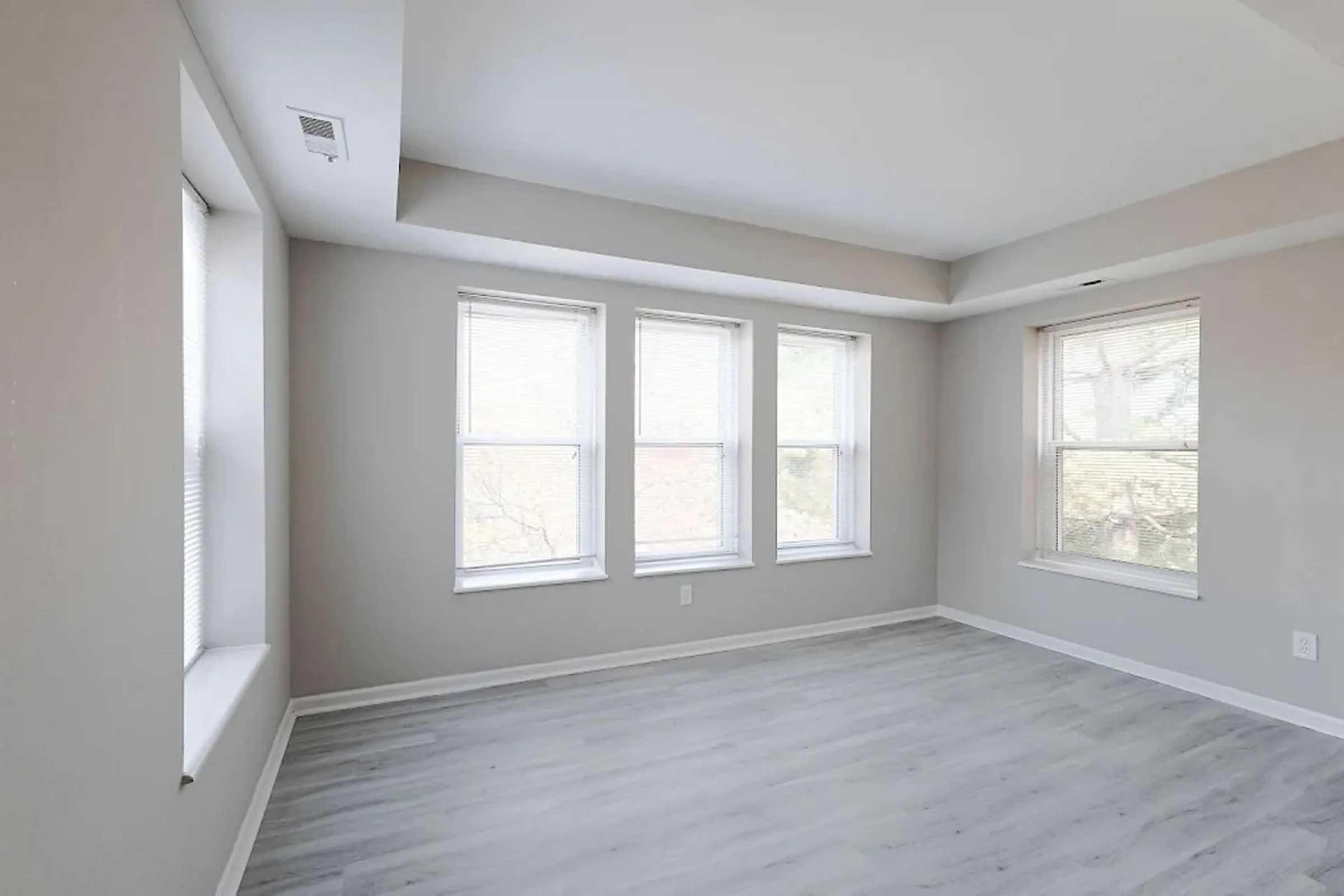 A bright, empty room with light gray walls and three windows allowing natural light. The flooring is light-colored and smooth, creating a spacious and airy atmosphere.