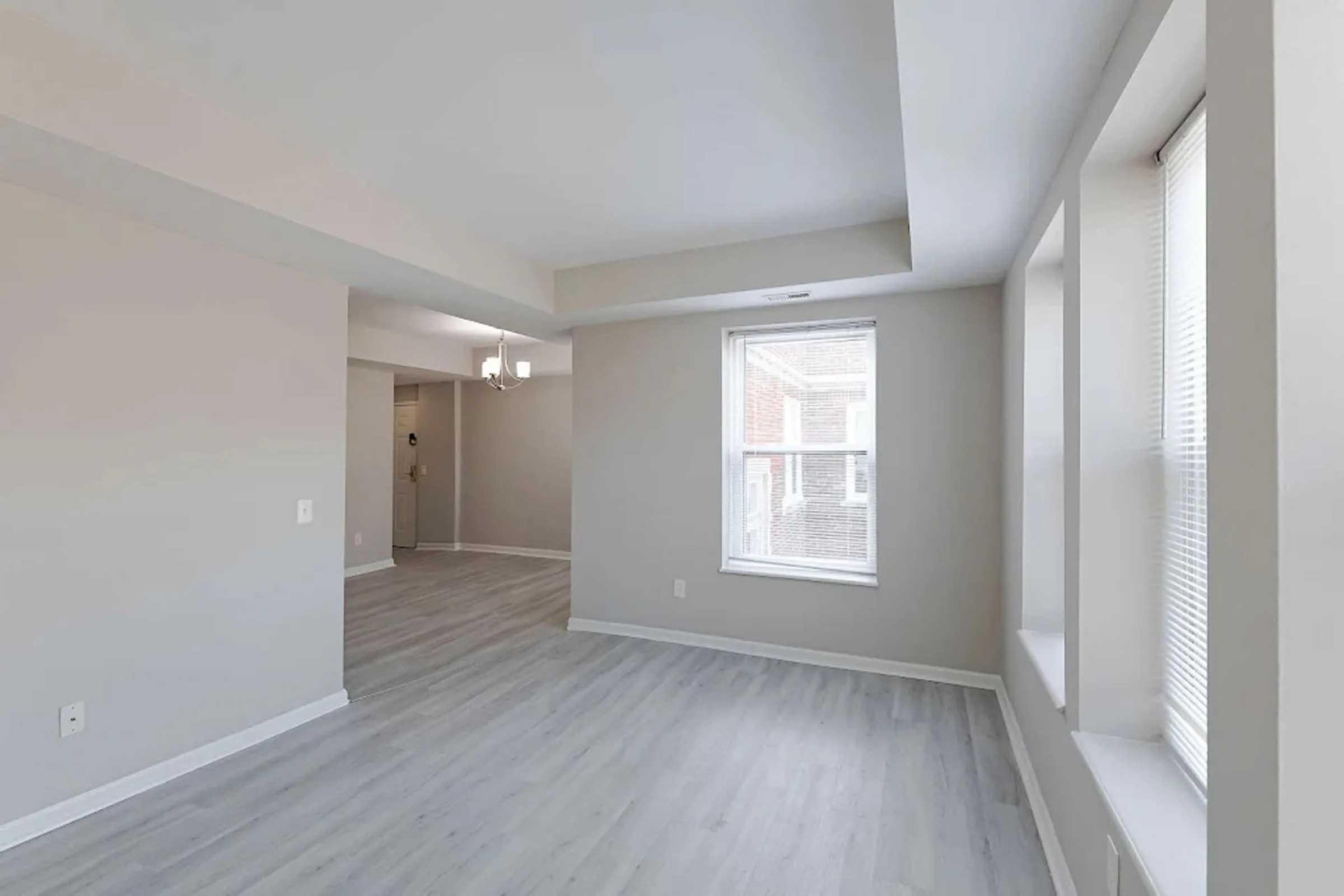 A spacious, light-filled living area with gray walls and white trim. Large windows provide natural light, and the floor features light-colored vinyl or laminate. The room is empty, showcasing modern design elements, including simple lighting fixtures and clean lines. Adjacent rooms are partially visible.