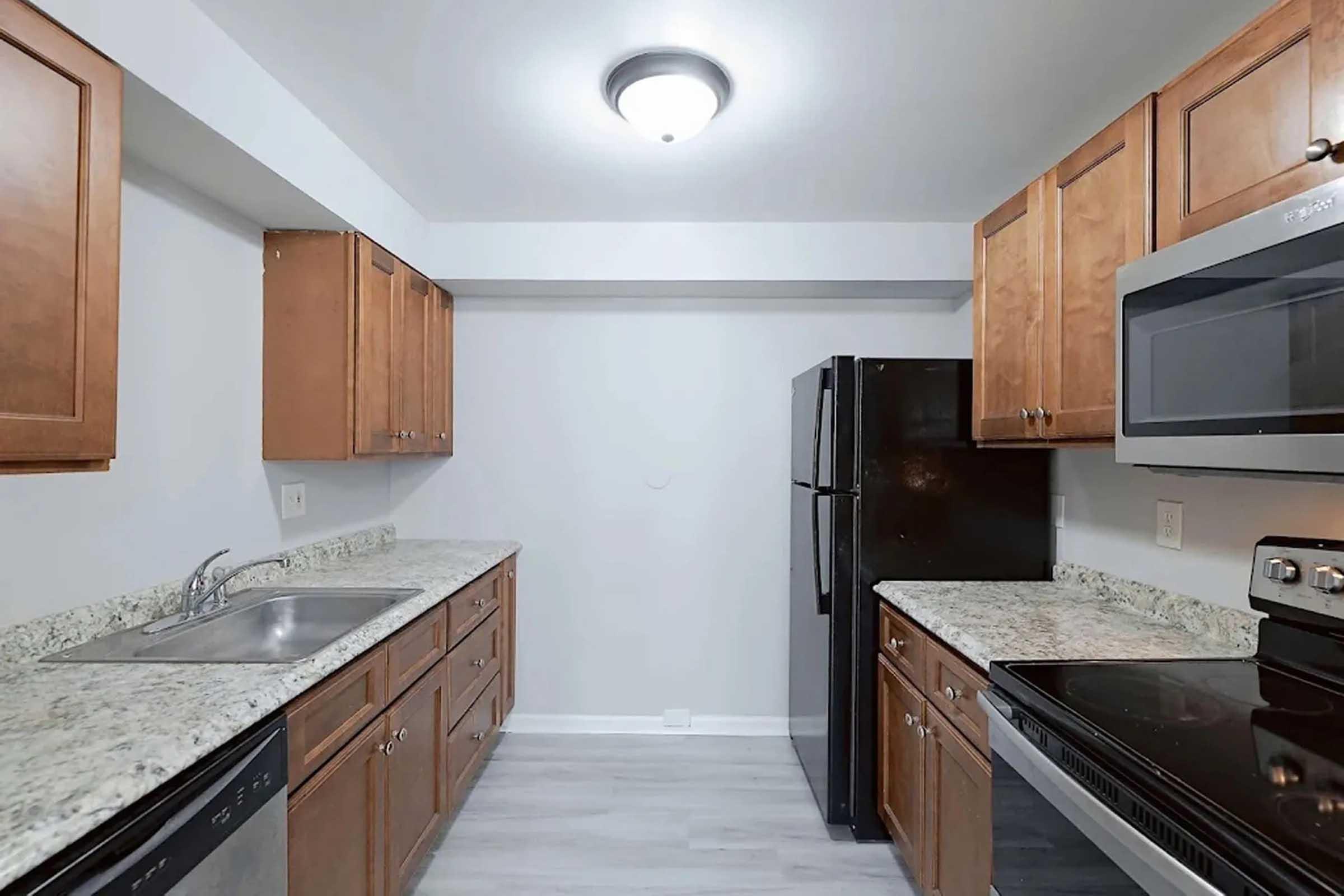 A modern kitchen featuring wooden cabinets, a granite countertop, and stainless steel appliances. It includes a double sink, a black refrigerator, a microwave, and an electric stove. The walls are painted light gray, and the flooring is a light-colored laminate. Bright overhead lighting enhances the space.