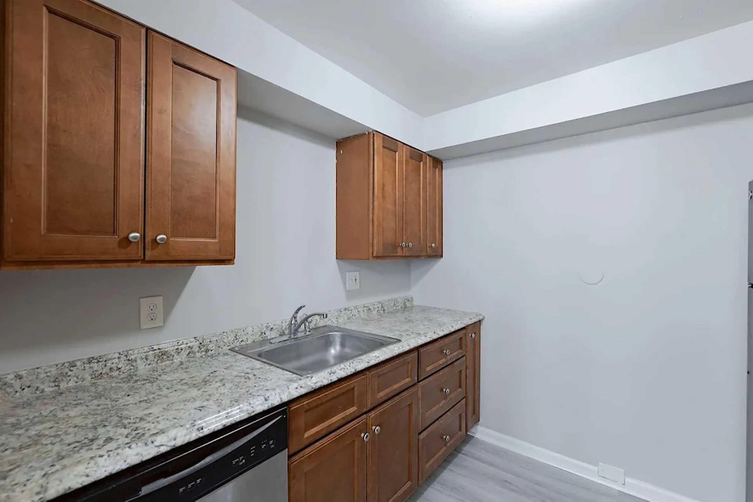 A modern kitchen featuring wooden cabinets, a granite countertop, and a stainless steel sink. The space includes a dishwasher and an open area with light-colored walls and gray flooring, providing a bright and clean appearance.