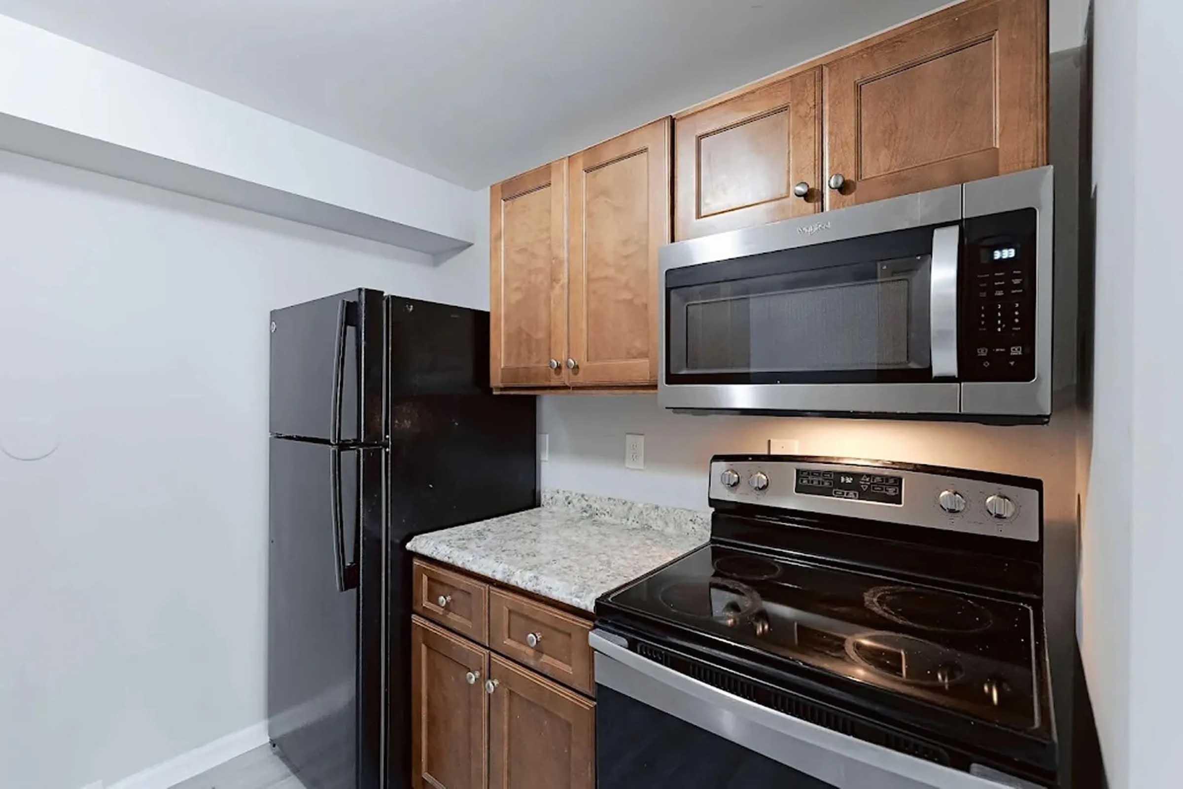 A modern kitchen featuring dark wood cabinets, a stainless steel microwave, and an electric stove. The countertop is made of speckled granite, and there is a black refrigerator alongside the cabinetry. The walls are painted in a light color, creating a clean and contemporary look.