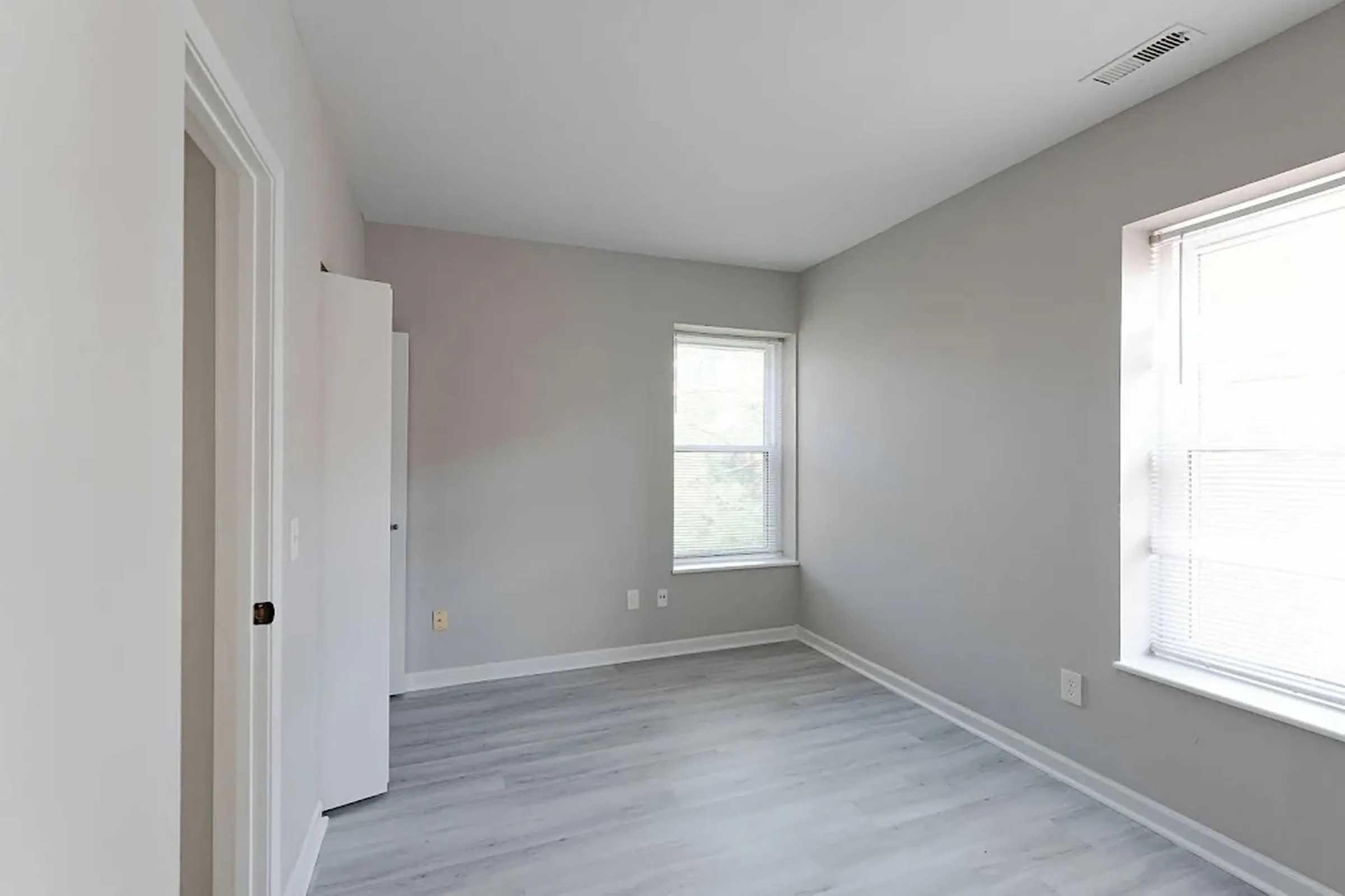 A bright, empty room featuring light gray walls and light-colored flooring. Two windows provide natural light, and there is a closet door on the left. The space is clean and minimalistic, ideal for various decorating options.