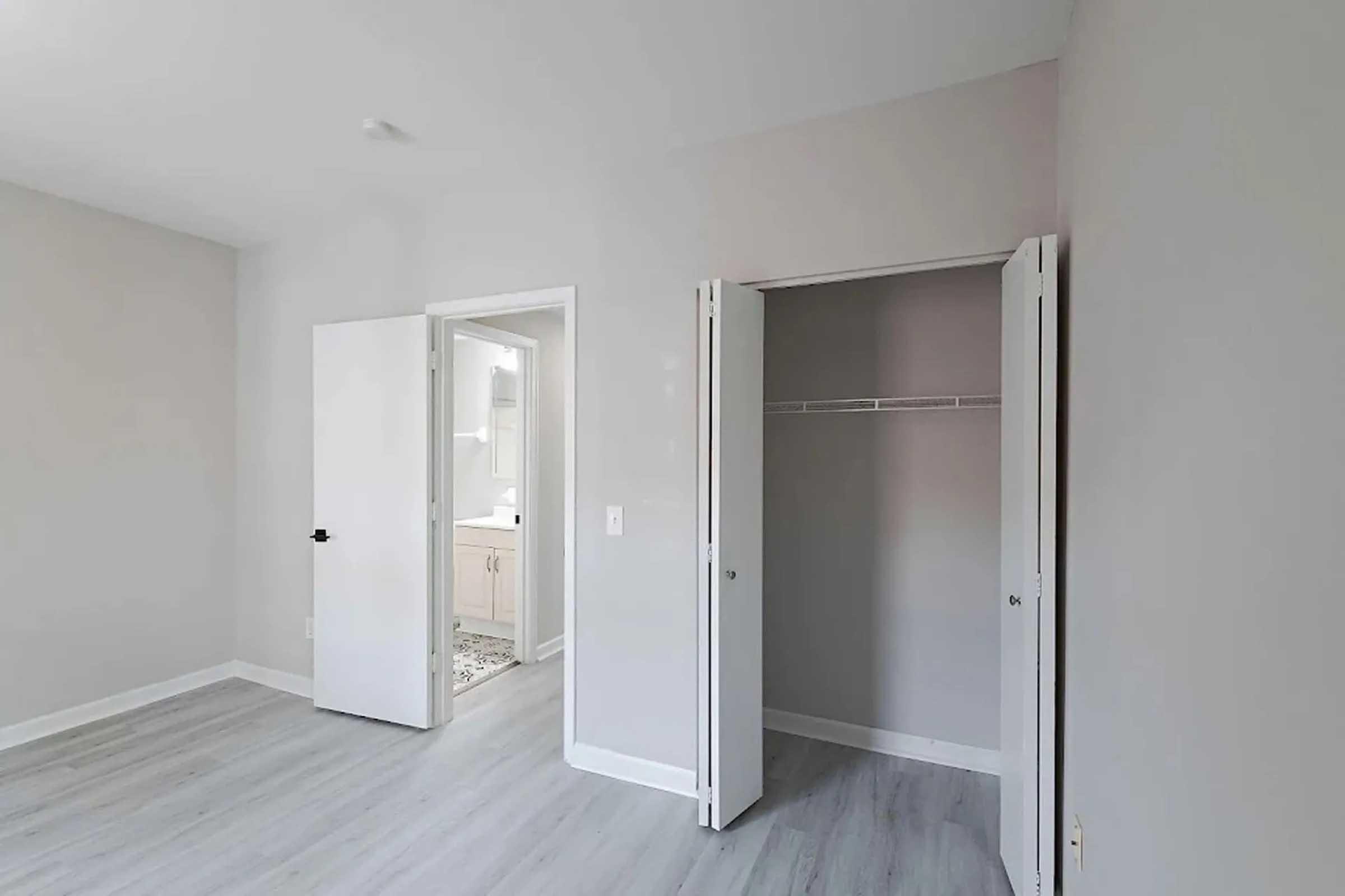 Empty room with light gray walls and light gray flooring. A closet with double doors is visible on the right, while a doorway leads to a bathroom or laundry area on the left. Natural light brightens the space.