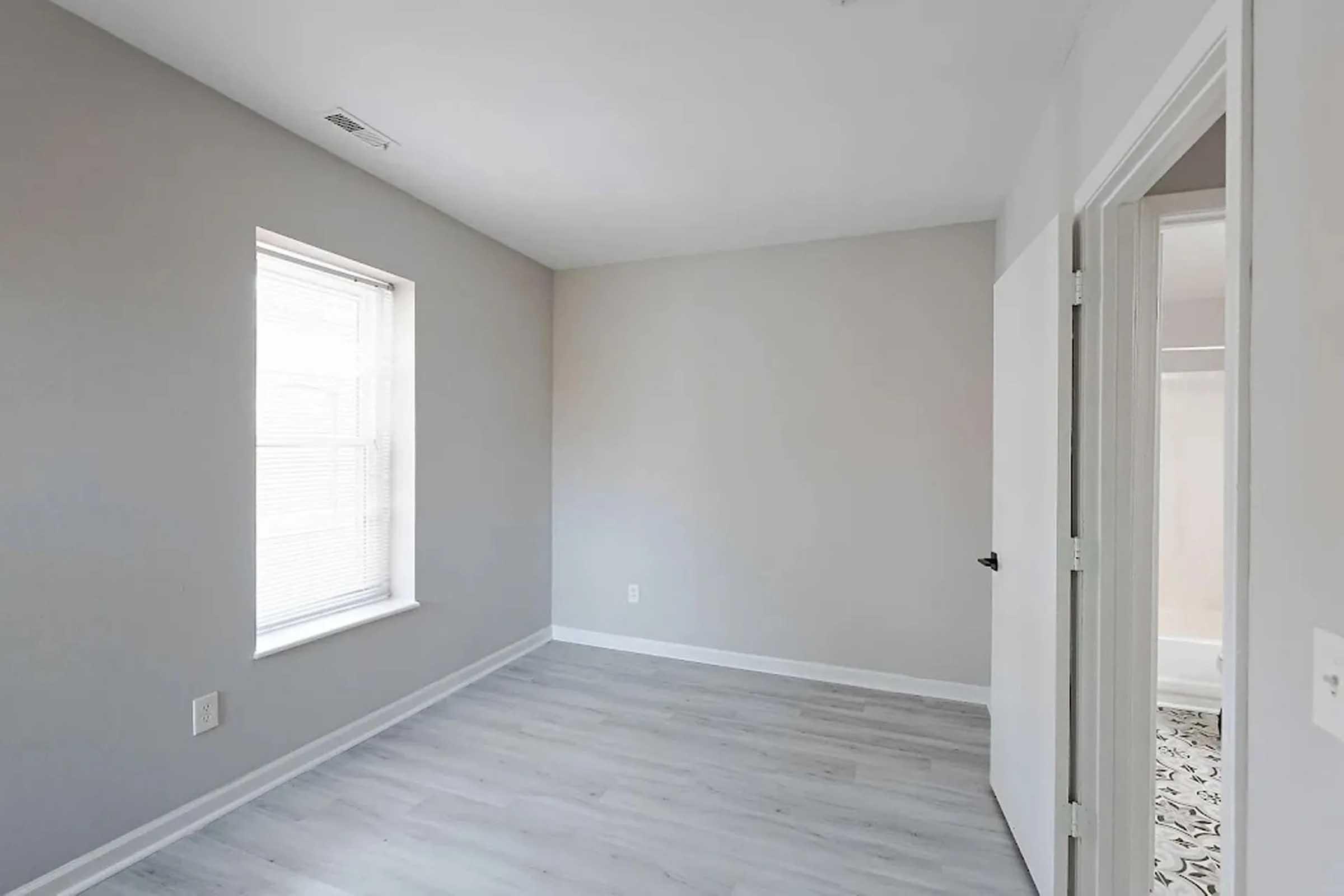 Empty room with light gray walls and light wood-style flooring. A window on one wall provides natural light, while an open door leads to another space. The overall atmosphere is bright and minimalist, suitable for various uses.