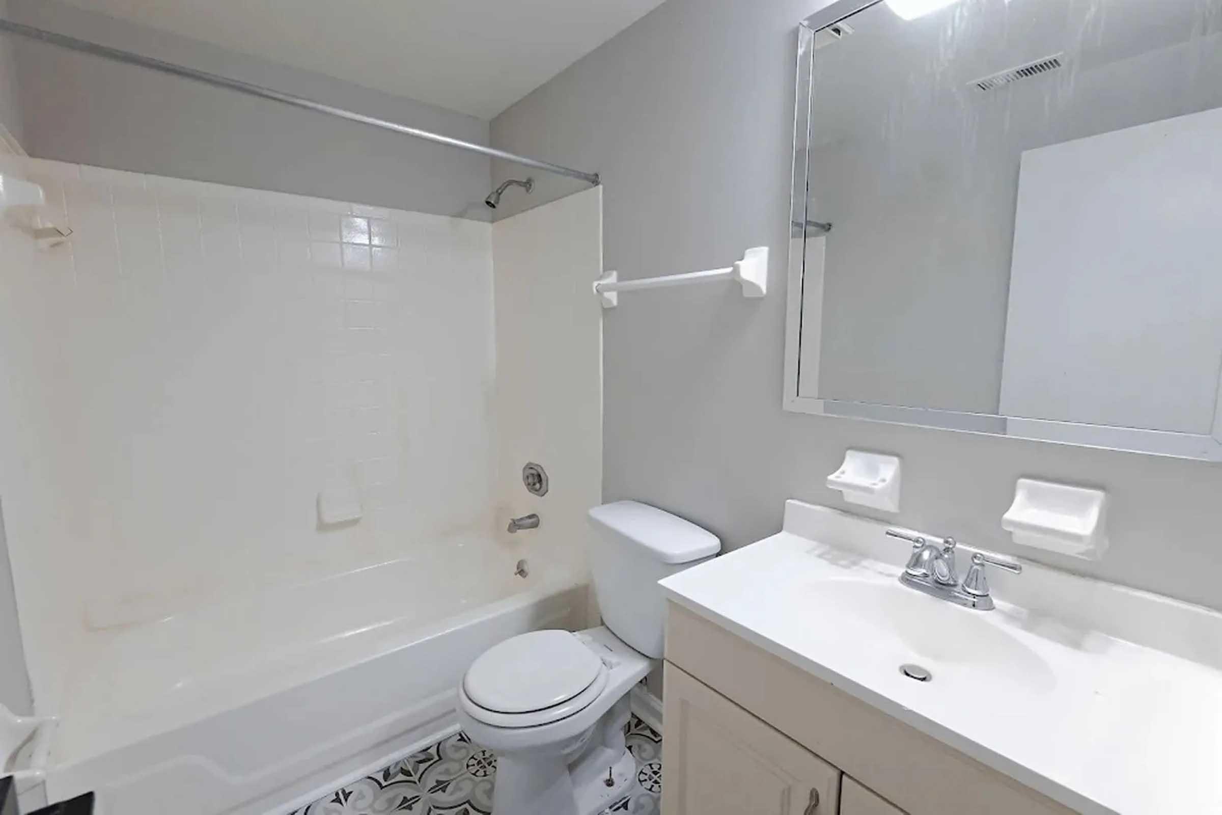 A clean, modern bathroom featuring a white bathtub with shower, a toilet, and a vanity with a sink. The walls are painted light grey, and the floor has a decorative black and white tile pattern. A large mirror is mounted above the sink, adding brightness to the space.