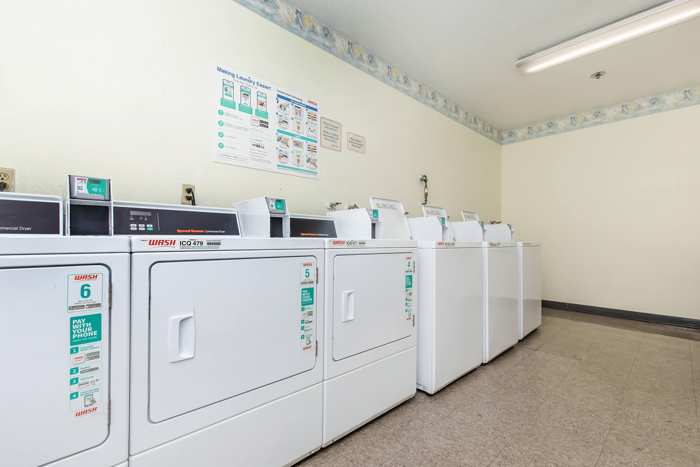 A laundry room containing several white washing machines and dryers lined up against a wall. The room has a light-colored, clean appearance with a bulletin board and signs on the wall providing laundry instructions. The floor is tiled, and there is overhead lighting.