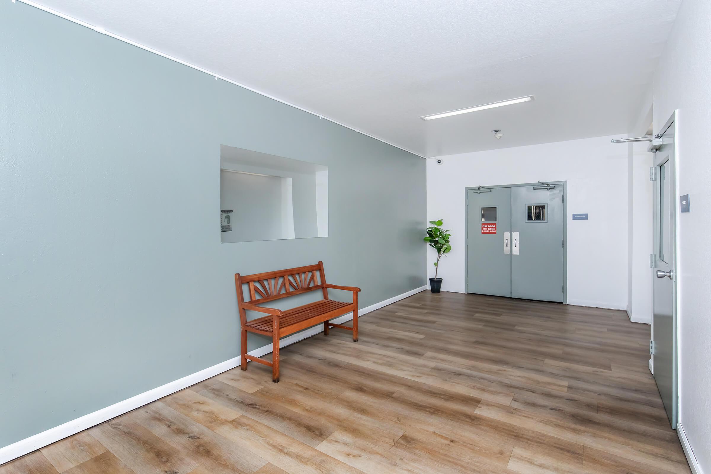 A spacious, well-lit hallway featuring a light gray wall and wooden flooring. There is a simple wooden bench against the wall and a potted plant nearby. Double doors are visible at the end of the hallway, leading to another area, and a large mirror is mounted on the wall.
