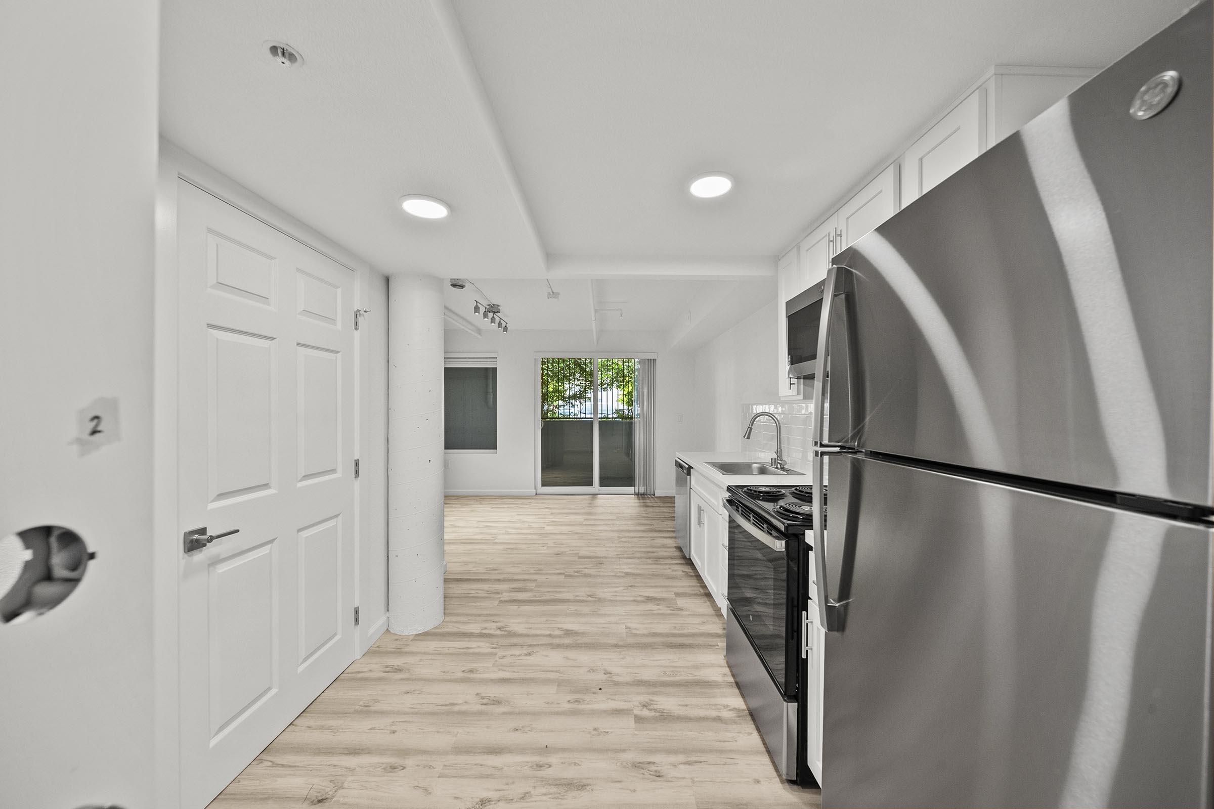 Modern kitchen featuring stainless steel appliances, including a refrigerator and gas stove. The space has white cabinetry and light wood flooring. There are two doors visible, and large windows allow natural light to fill the room, creating a bright and airy atmosphere.