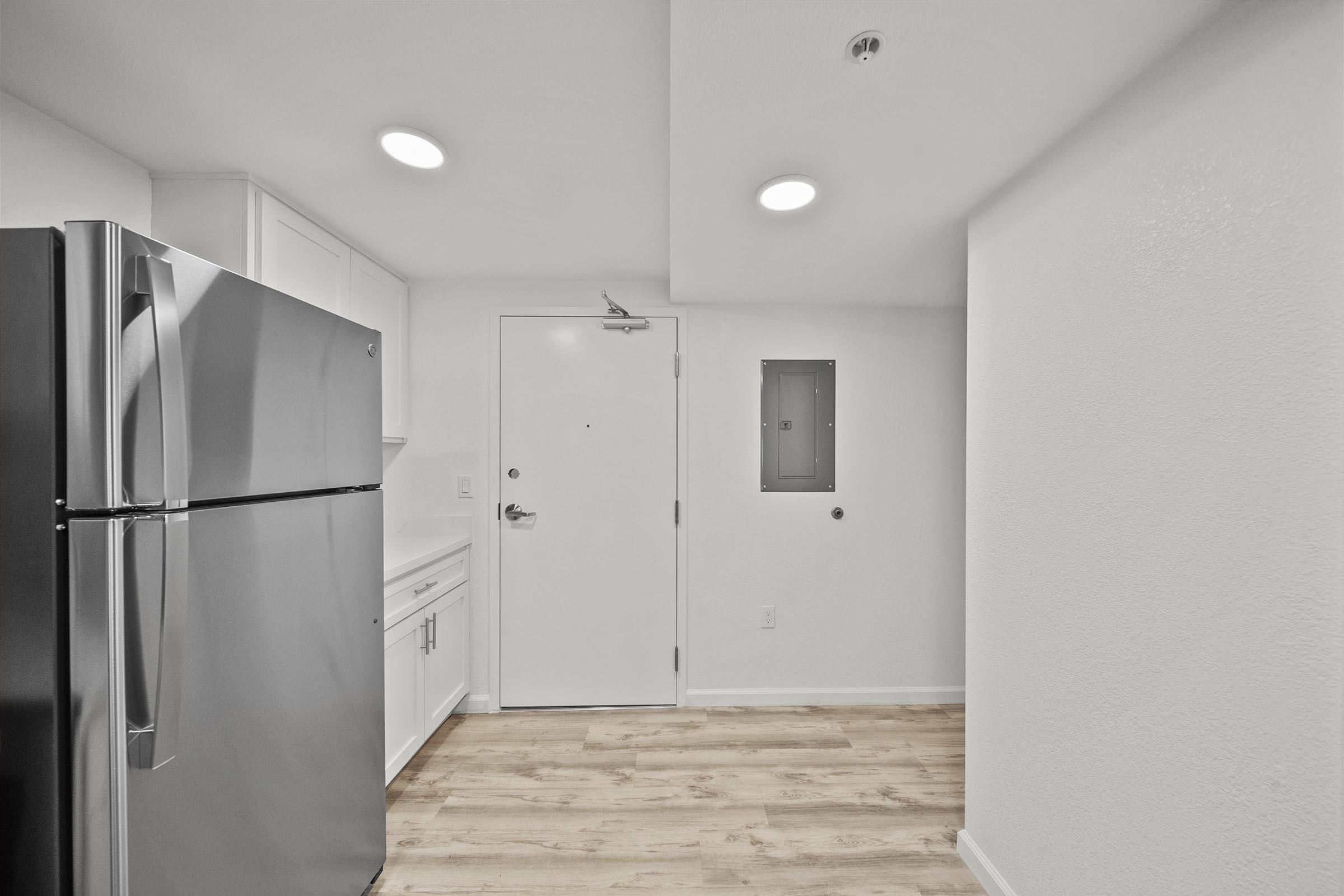 A modern kitchen area featuring a stainless steel refrigerator, white cabinets, and light-colored walls. The space includes a door leading outside, a small electrical panel on the wall, and hardwood-style flooring, creating a clean and bright atmosphere.