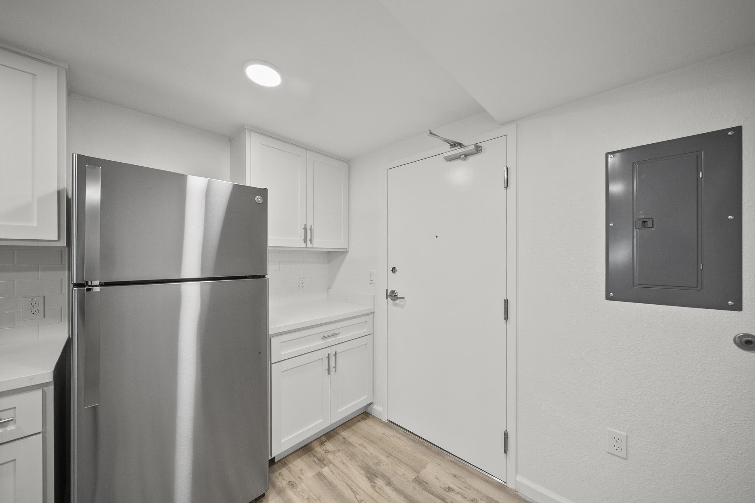 A modern kitchen with white cabinetry, a stainless steel refrigerator, and a solid door leading outside. The space features light-colored walls, a beige wood floor, and an electrical panel mounted on the wall near the entrance. Bright lighting enhances the clean and minimalist design.