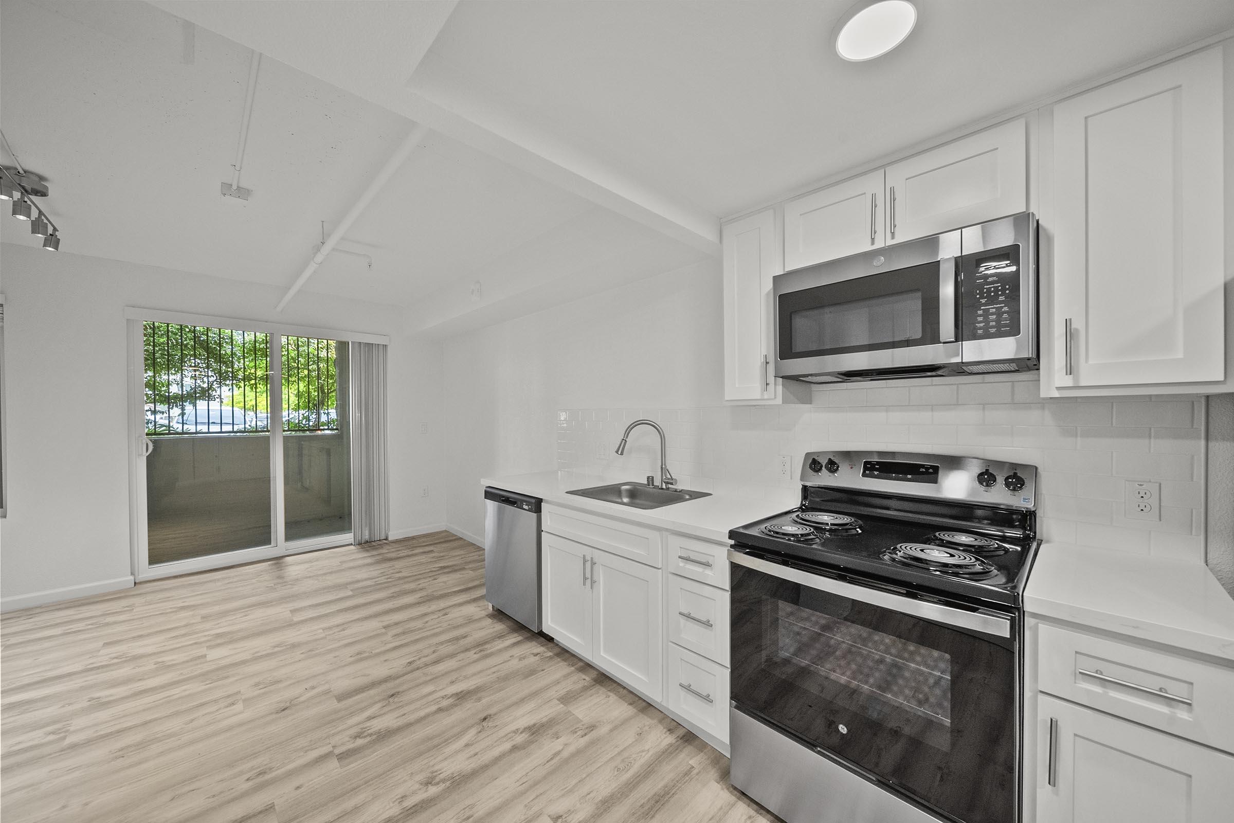 A modern kitchen featuring white cabinets, stainless steel appliances including a microwave, stove, and dishwasher, with a spacious countertop. A large window allows natural light to fill the room, complementing the light wood flooring and creating a bright, inviting atmosphere. 