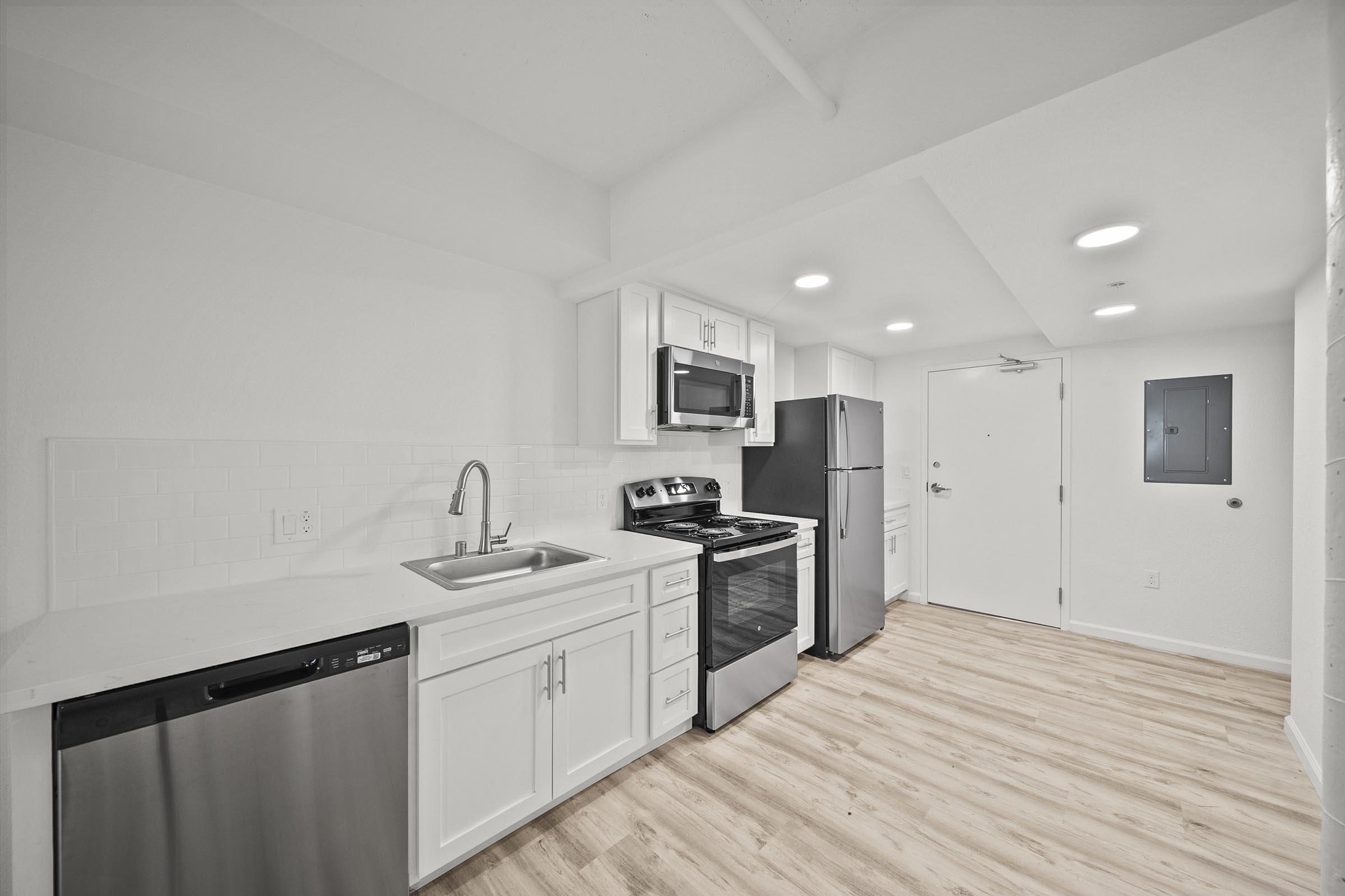 Modern kitchen featuring white cabinetry, stainless steel appliances including a refrigerator, oven, microwave, and dishwasher. The countertops are light-colored, and the flooring is a light wood finish. Bright lighting and a door leading to another area are visible.