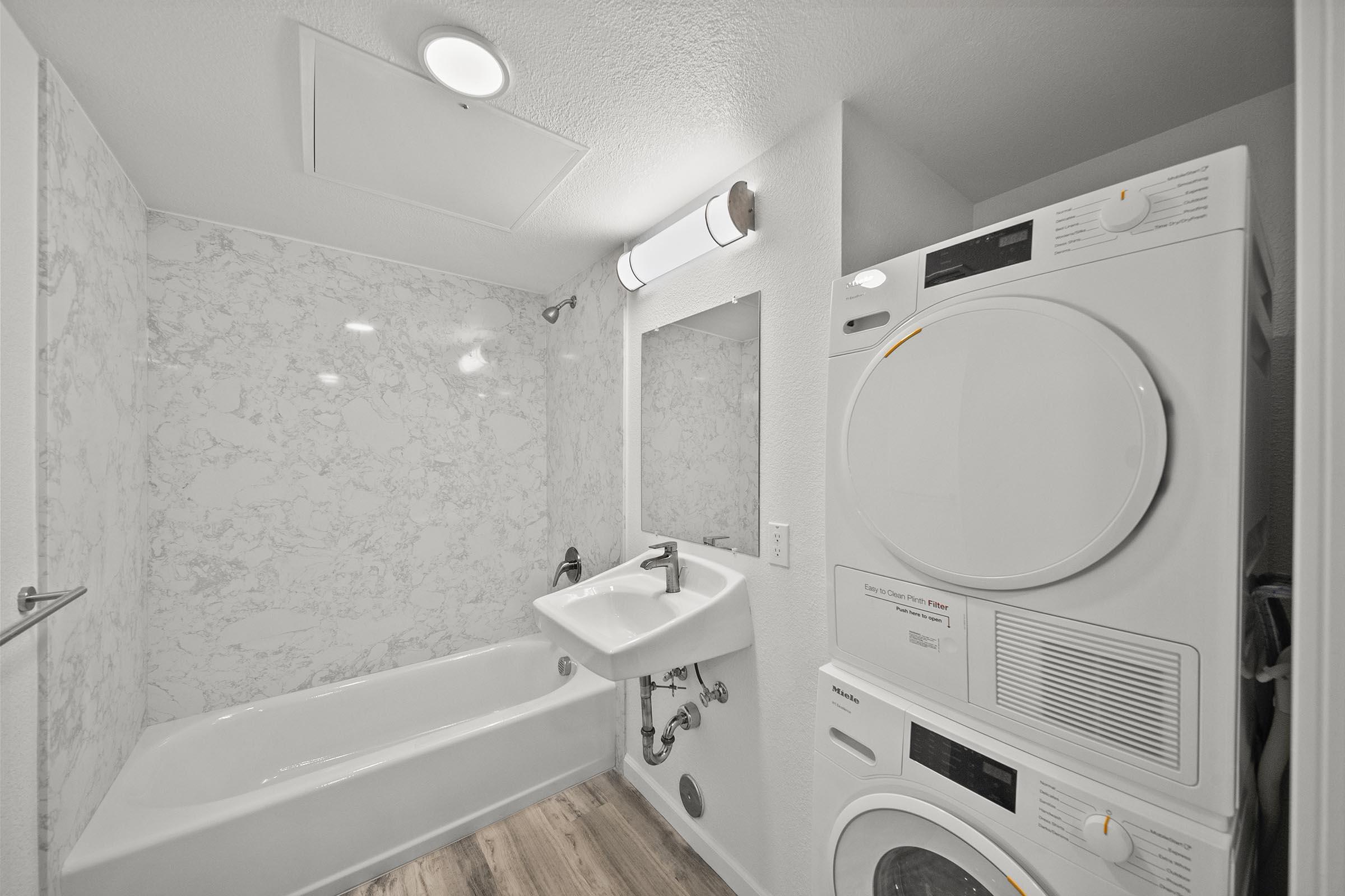 A modern bathroom featuring a white tub with a marble backsplash, a sink, a large mirror, and a stacked washer and dryer unit. The space is well-lit with a ceiling light and has a minimalist design with light wood flooring.