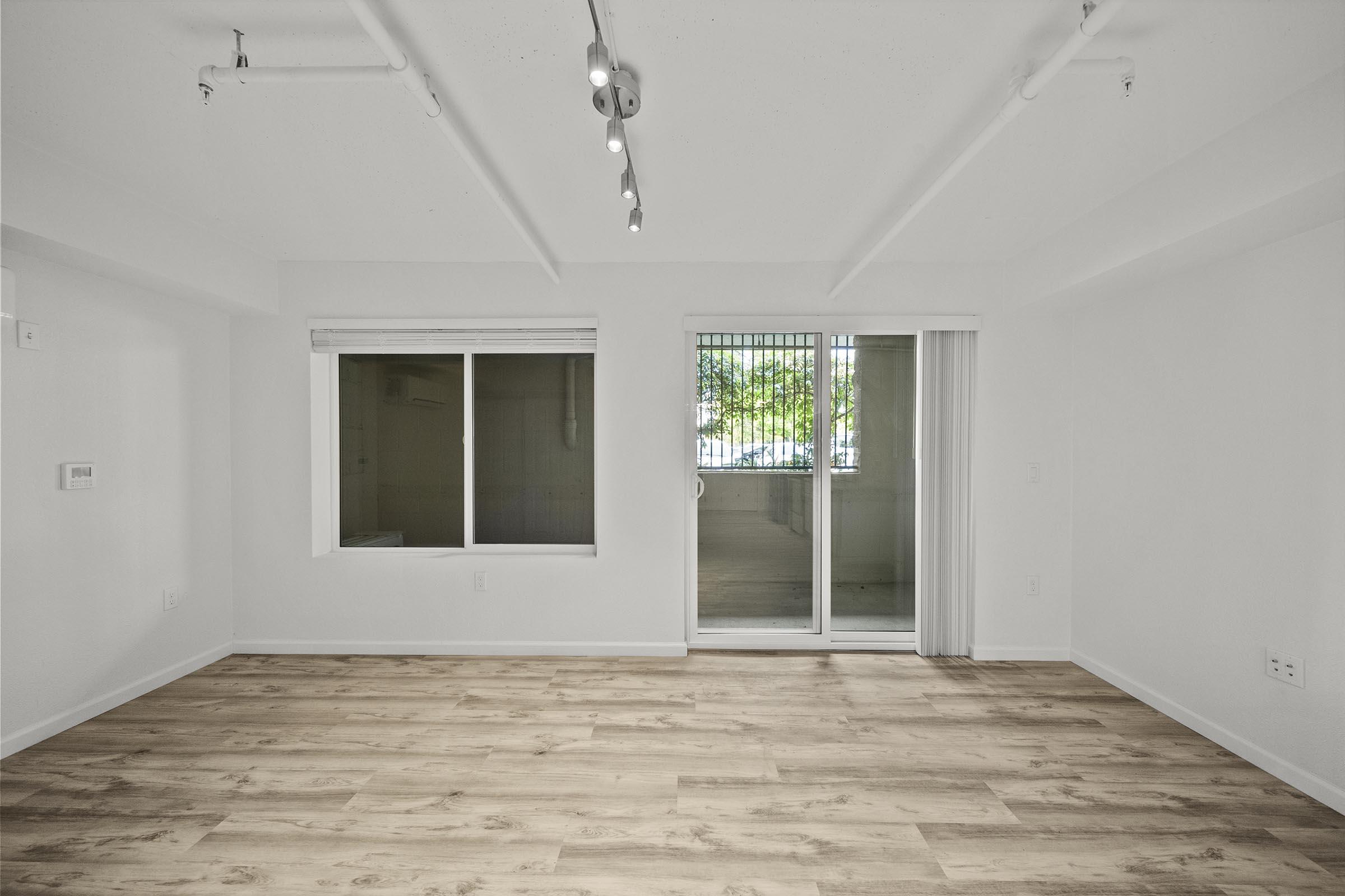 Empty modern living room with light-colored walls and wooden flooring. A large window on the left side offers natural light, while a sliding glass door leads to a balcony or patio. There are exposed beams on the ceiling and track lighting overhead, creating a bright and spacious atmosphere.