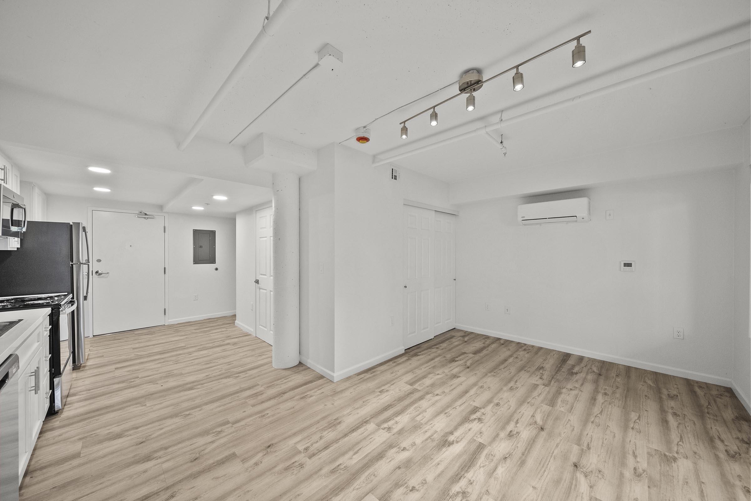 A modern, empty apartment interior featuring a kitchen on the left with stainless steel appliances and a living area on the right. The space has light-colored wooden flooring, white walls, and recessed lighting, creating a bright and airy atmosphere. A door leads to the hallway, and there's an air conditioning unit on the wall.