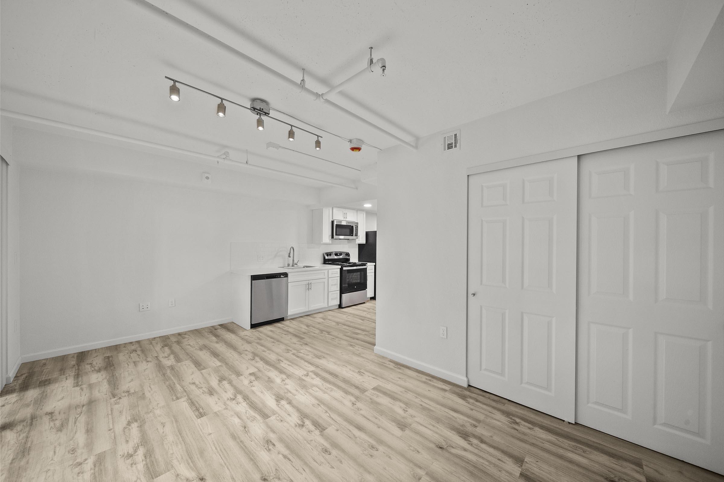 A modern, open-concept living space featuring light wood flooring, white walls, and recessed lighting. The kitchen is equipped with black and stainless steel appliances, including a microwave and stove. There are dual sliding closet doors on the right, enhancing the room's functionality and minimalistic design.