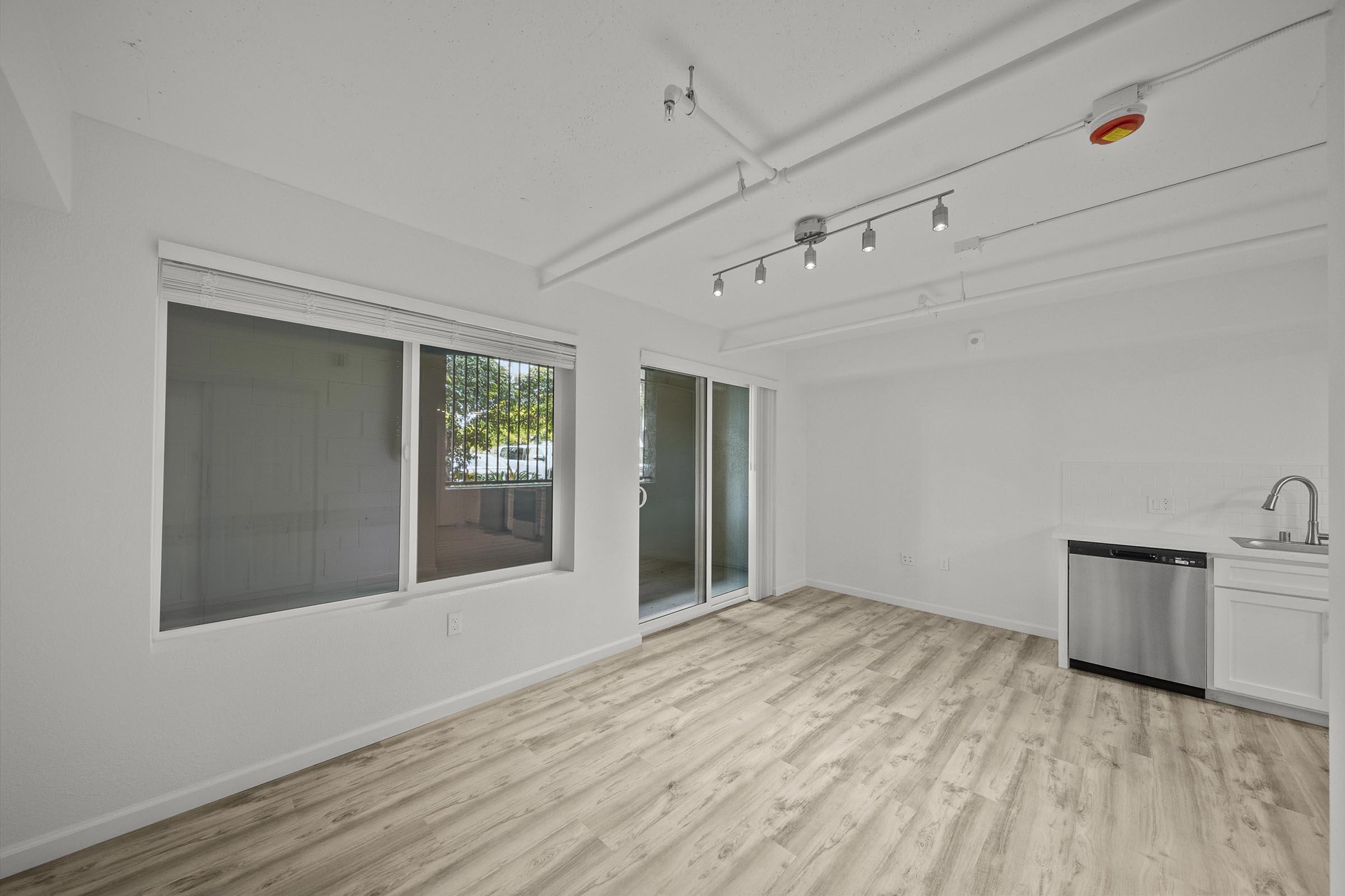 A bright, empty living space with white walls, large windows, and sliding glass doors that open to a patio. The floor is light-colored wood, and there is a kitchen area with a dishwasher and sink in the corner. The ceiling features exposed ductwork and track lighting.