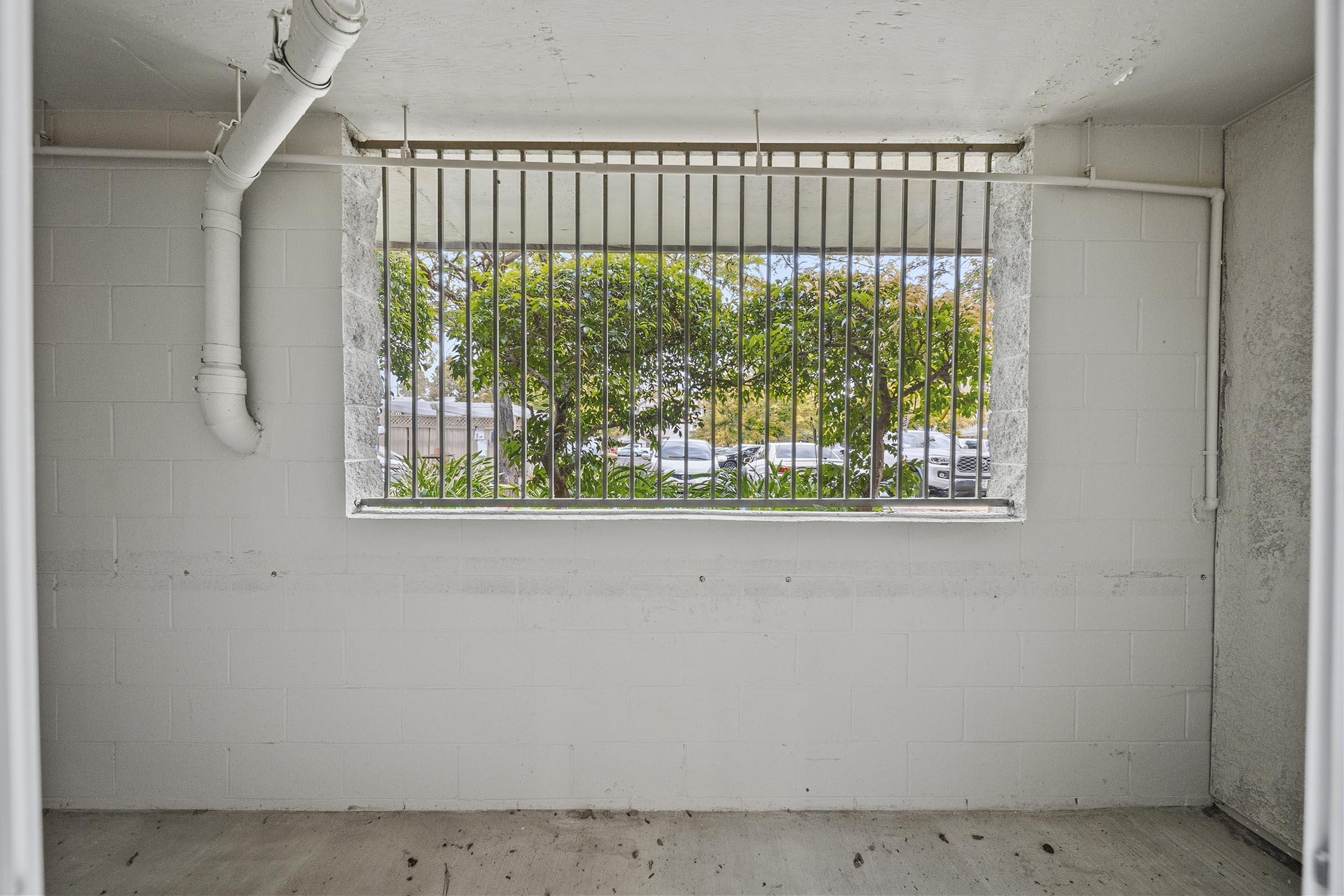 A view from inside a room looking out through a barred window. The window frames greenery and parked cars outside. The walls are bare and painted white, and the floor appears to be concrete. Natural light enters the space, highlighting the window bars.