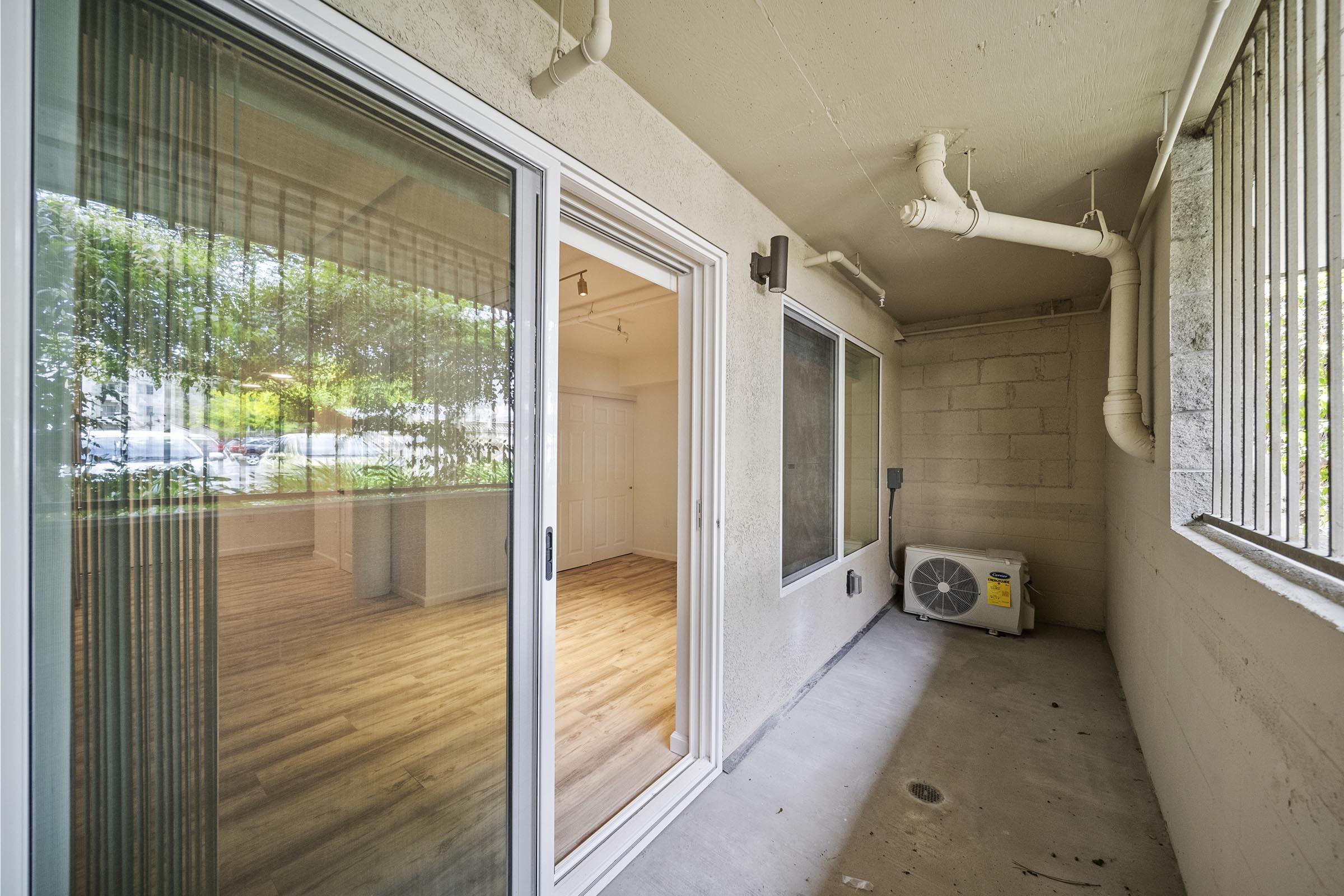 A covered patio area with sliding glass doors leading to an interior space. The floor is concrete, and there is an air conditioning unit visible. Natural light filters in, highlighting the wooden flooring inside the room.