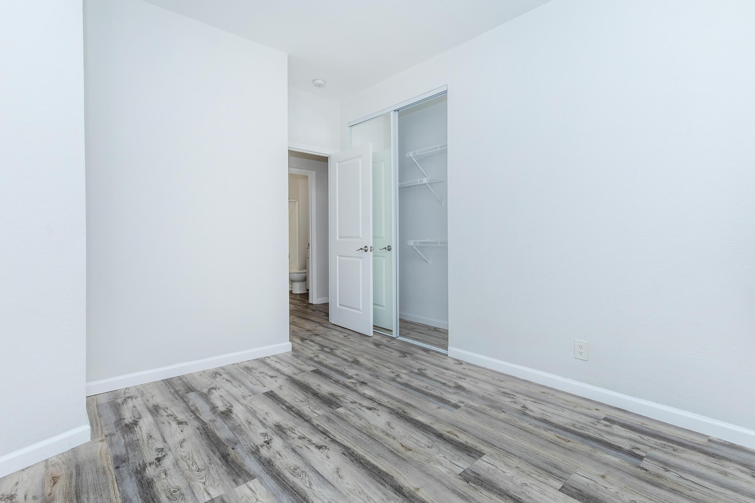 A clean, empty room featuring light-colored walls and a wooden floor. A partially open closet with sliding doors is visible on the left, and a doorway leads to another space in the background. The overall aesthetic is simple and minimalistic, providing a bright and spacious feel.