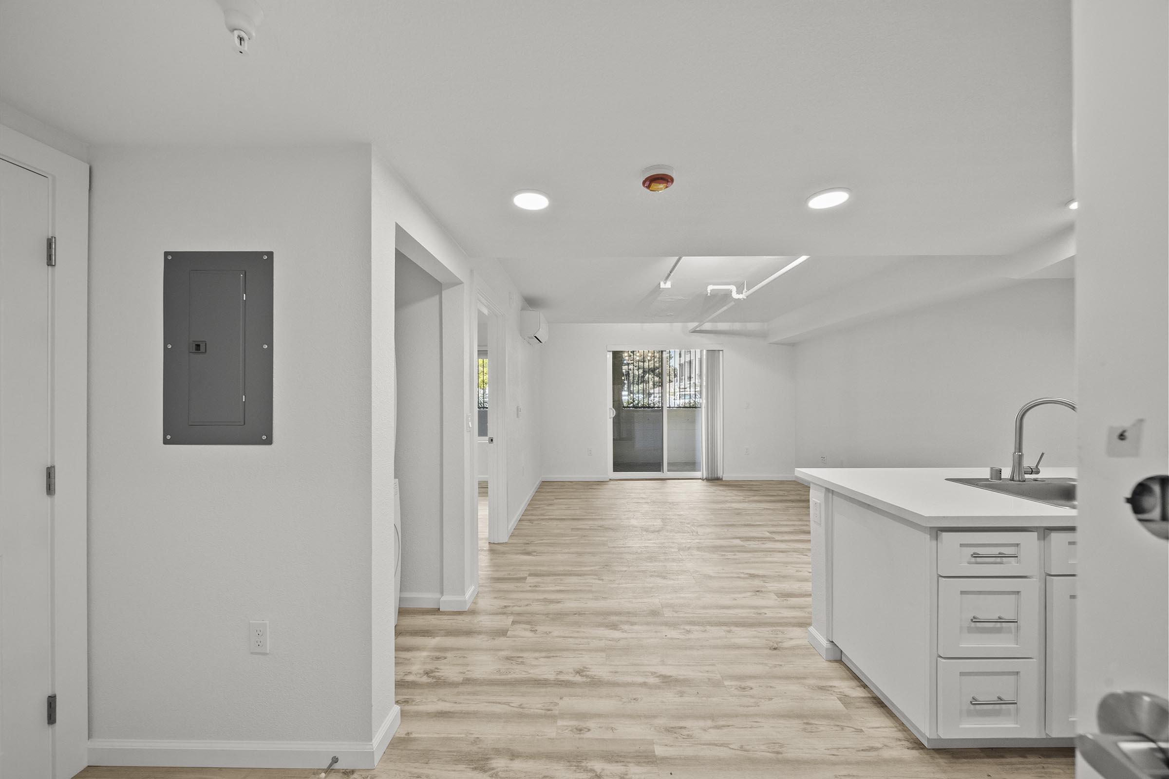 A bright, modern apartment interior showcasing an open layout. The view includes a kitchen island with white cabinetry, light hardwood flooring, and large windows allowing natural light. The space appears clean and inviting, with neutral-colored walls and minimalistic design elements.