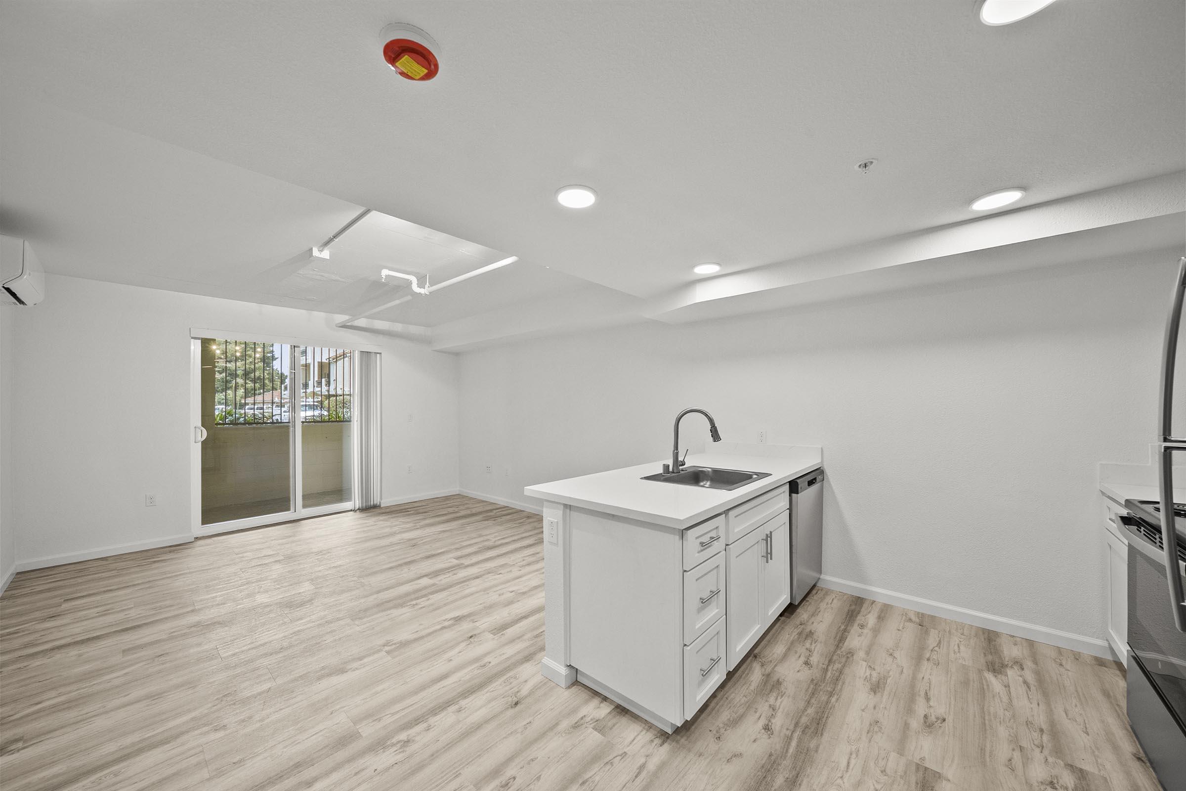 Bright, modern open-concept kitchen and living area with white cabinetry, a stainless steel sink, and appliances. The space features light wood flooring, recessed lighting, and a sliding glass door leading to an outdoor area. The walls are painted in a neutral tone, creating a spacious and inviting atmosphere.