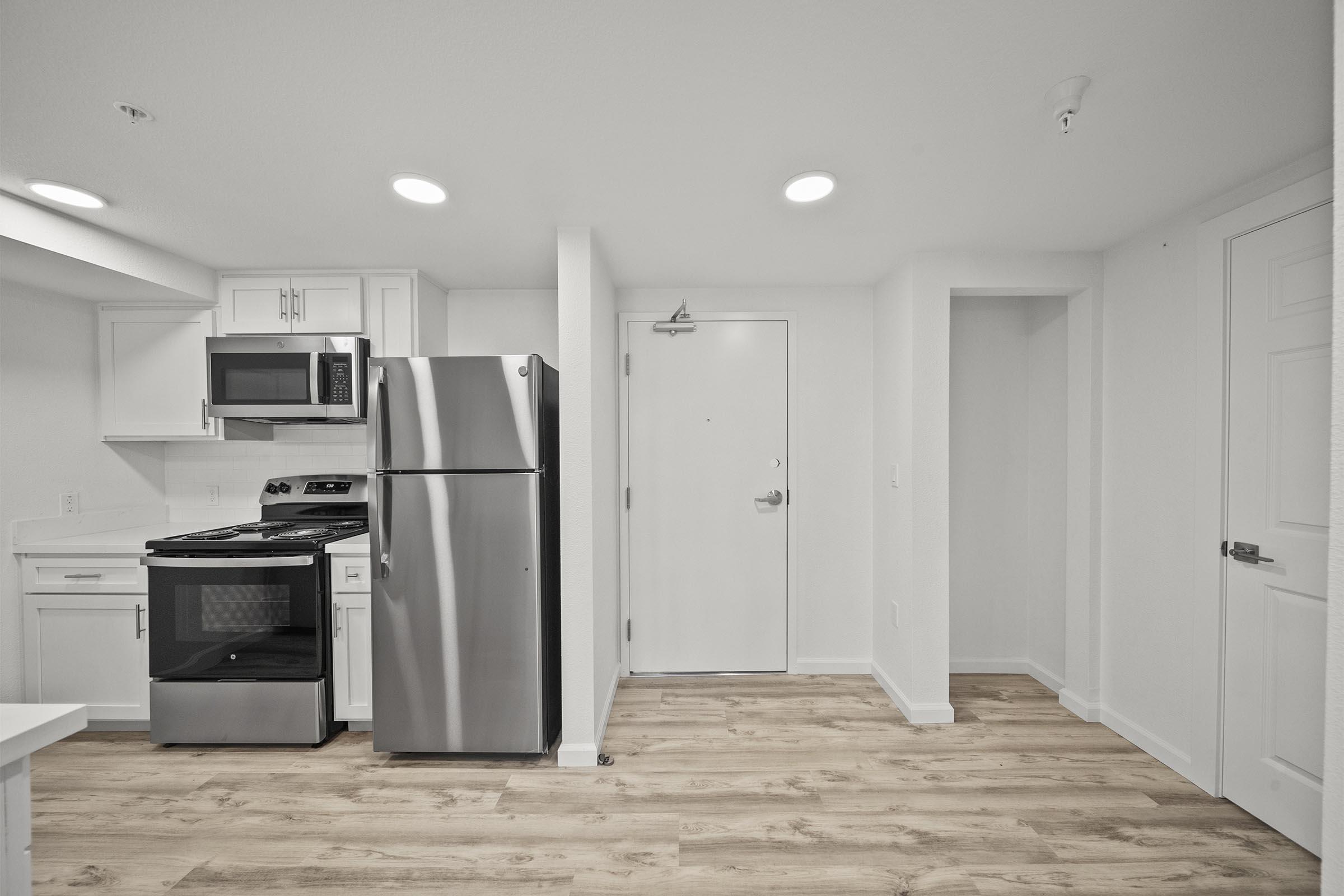 A modern kitchen area featuring a stainless steel refrigerator and microwave, a stove with an oven, and white cabinetry. The space has light-colored hardwood flooring and bright overhead lighting, with a door leading outside visible in the background. The overall design is clean and minimalistic.
