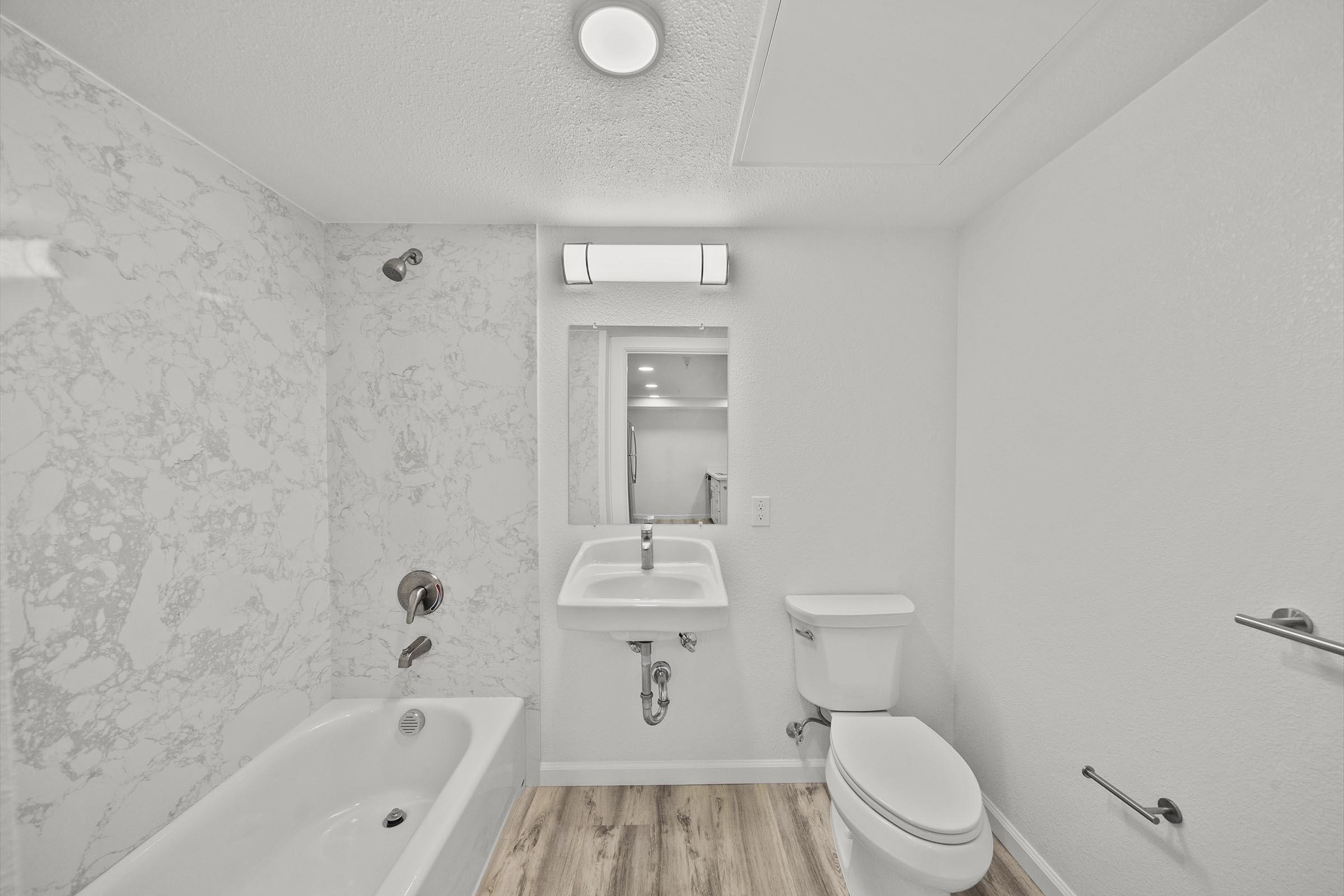 A modern bathroom featuring a white bathtub with a marble surround, a wall-mounted sink with a mirror above it, and a toilet. The walls are mostly white, and the flooring is light wood. A ceiling light fixture illuminates the space, creating a bright and clean ambiance.