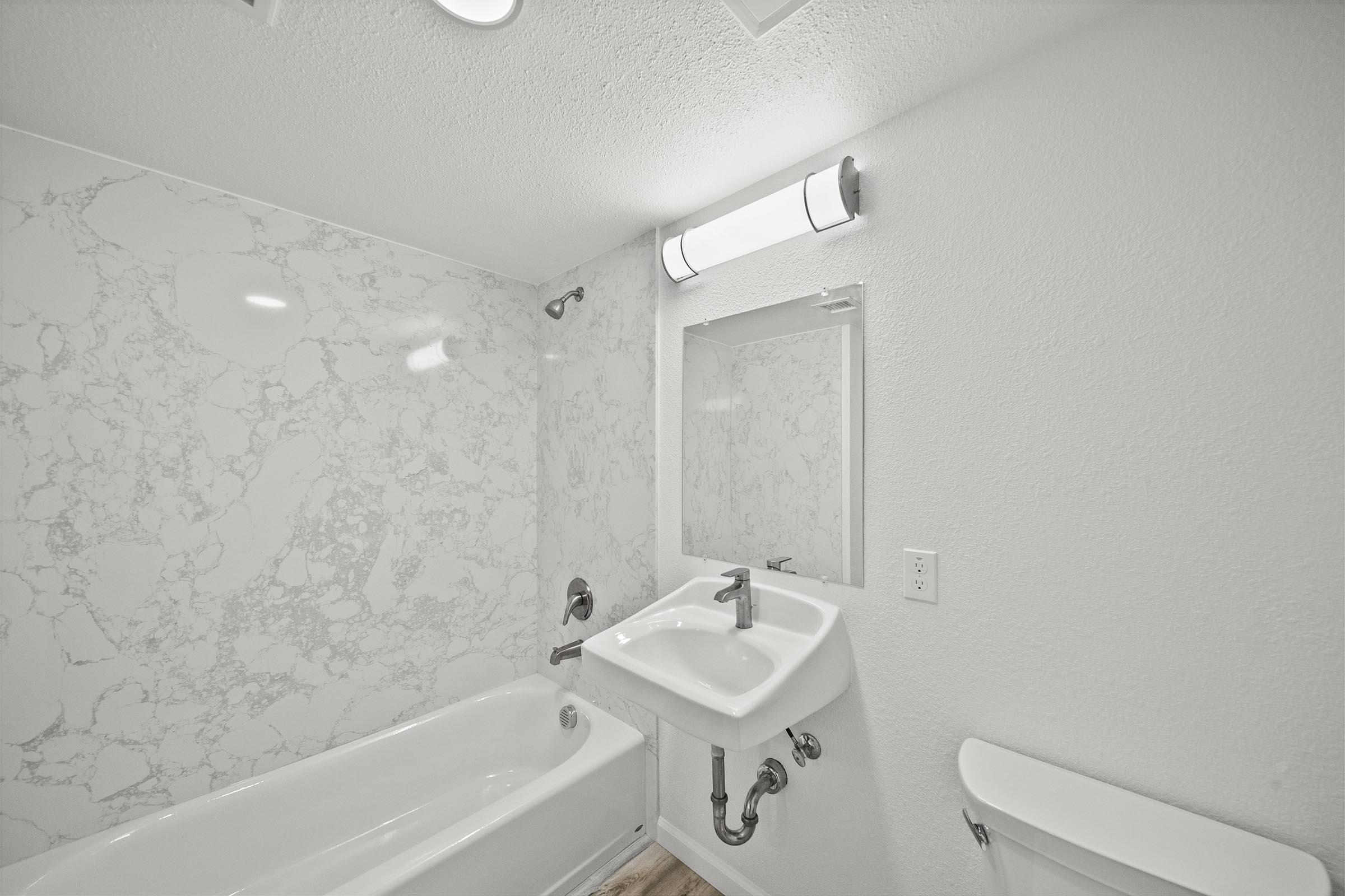 A modern bathroom featuring a white bathtub, a wall-mounted sink, and a large mirror. The walls are adorned with a light-colored marbled pattern, and there is a bright ceiling light above. A clean, minimalist design is highlighted by light wood flooring.