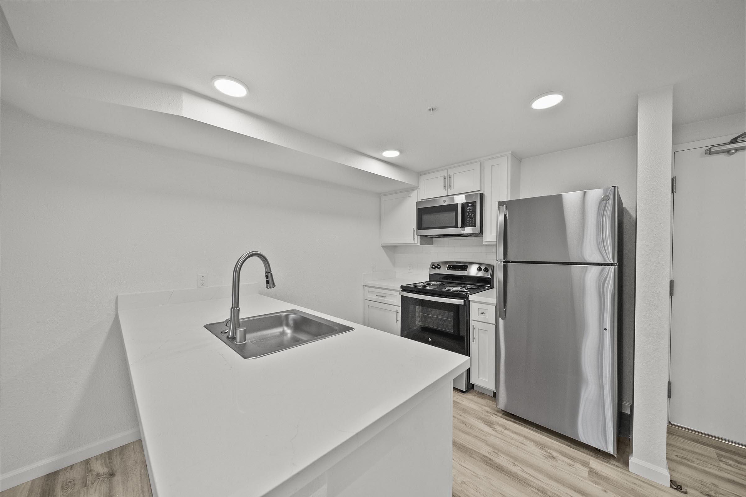 Modern kitchen with white cabinetry, stainless steel appliances including a microwave, oven, and refrigerator. A large island with a sink is in the foreground, and the space features light wood flooring and recessed lighting for a bright, open feel.