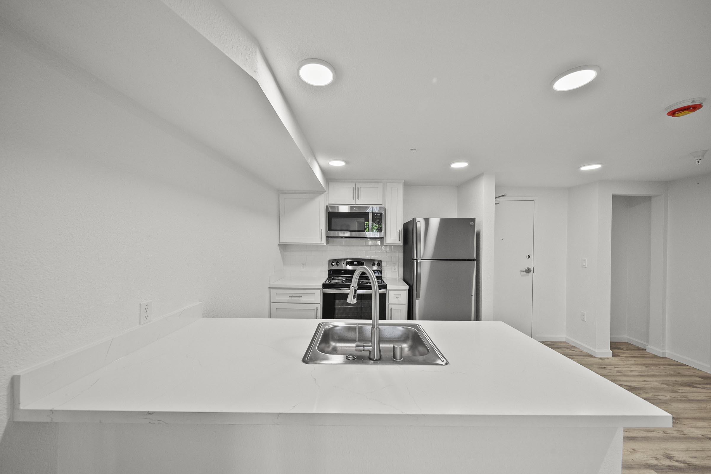 Modern kitchen with white cabinetry, stainless steel appliances including a fridge and stove, and a center island featuring a sink. The walls are painted white, and the flooring is light wood. Bright lighting from recessed fixtures enhances the spacious feel of the room.