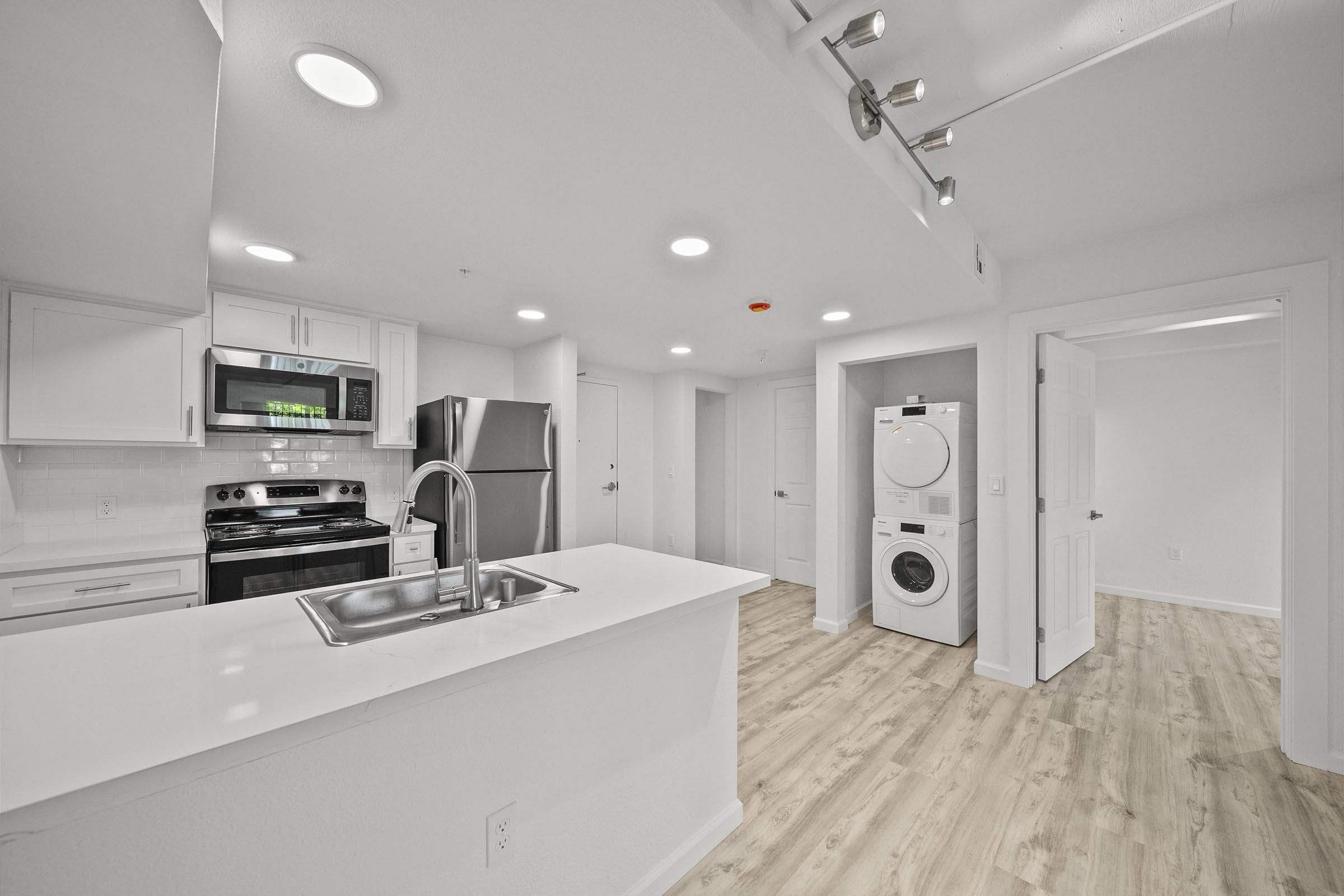 Modern kitchen and laundry area with stainless steel appliances, including a stove and refrigerator. The open layout features a white countertop and cabinetry, with a washer and dryer in a dedicated space. The flooring is light wood, and the walls are painted white, creating a bright and airy atmosphere.