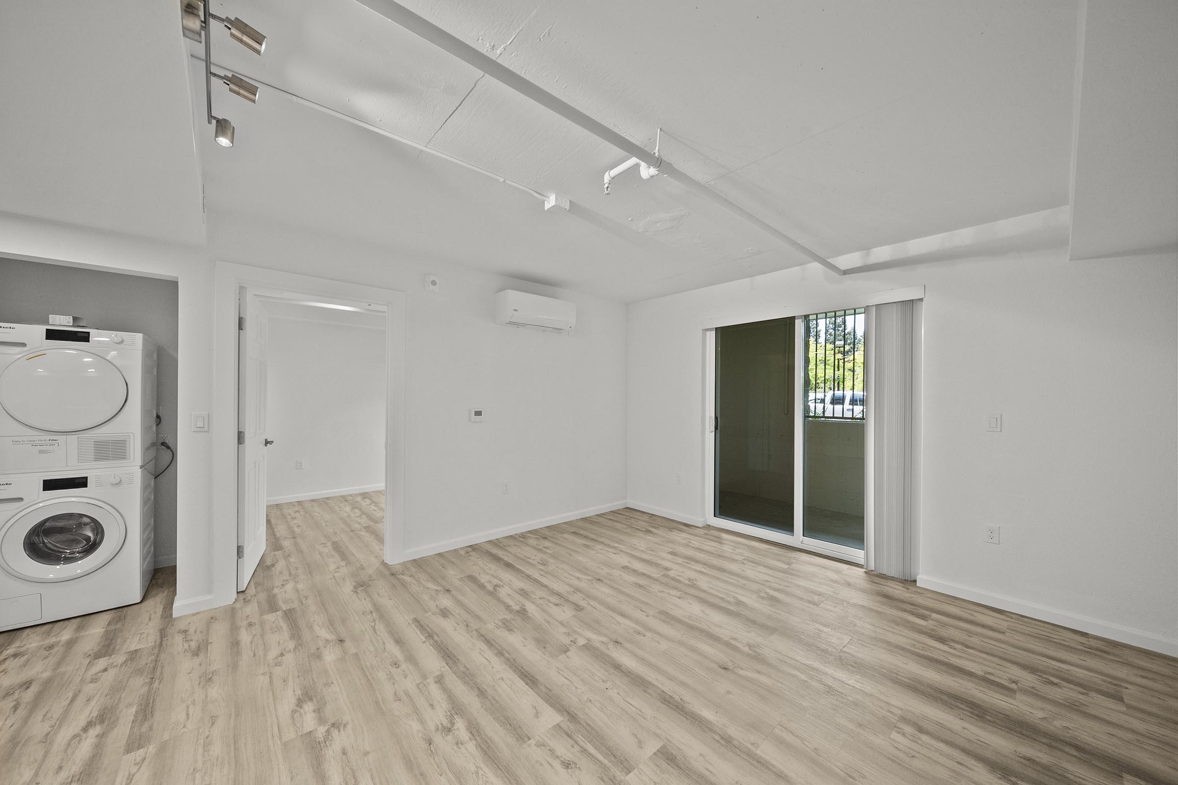 A bright, modern room with light wood flooring, featuring a wall-mounted air conditioner and a sliding glass door leading to an outdoor area. On the left, there is a stacked washer and dryer unit. The walls are painted white, contributing to a clean and spacious atmosphere.