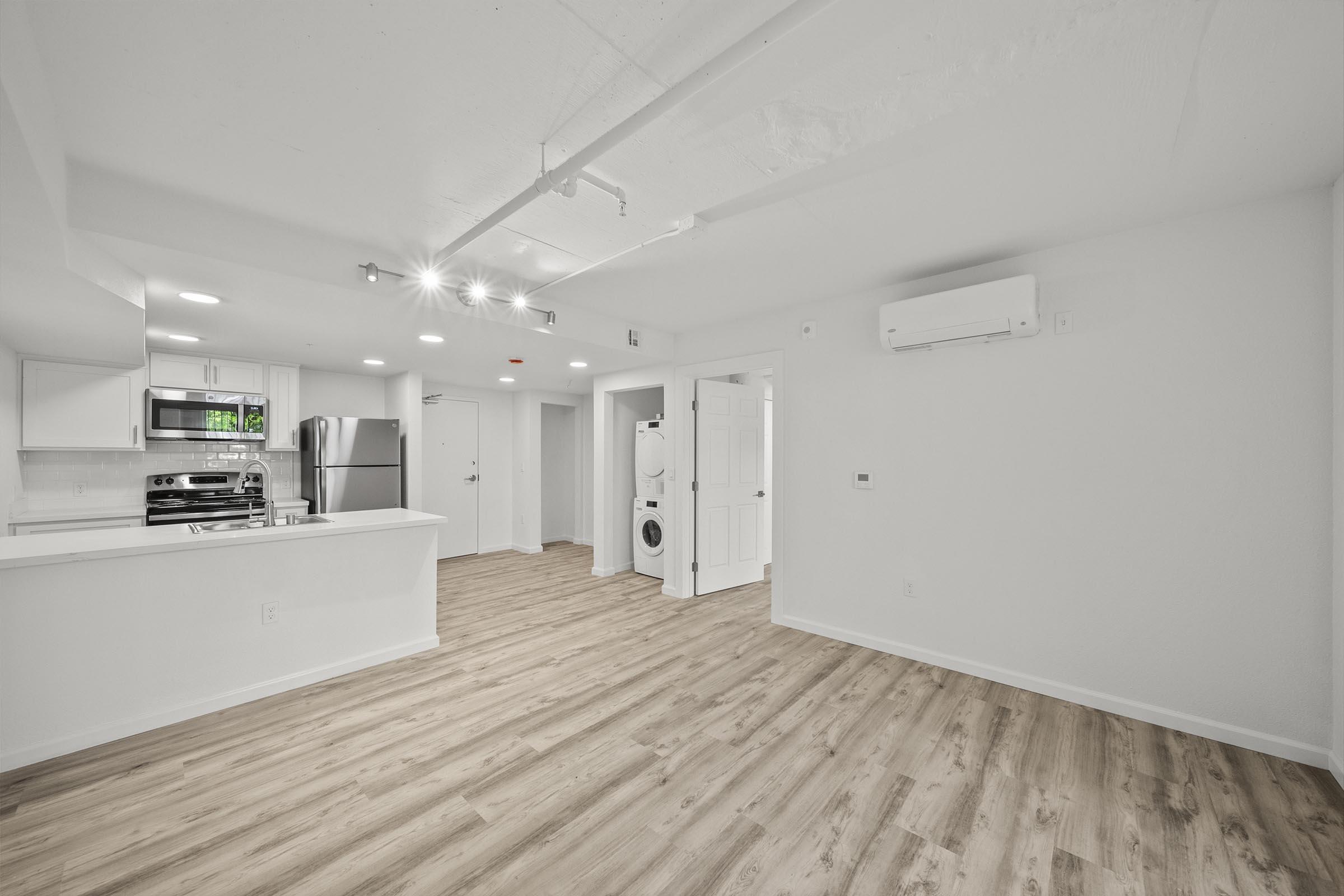 A spacious, modern interior of an apartment featuring light wood flooring, recessed lighting, and an open kitchen with stainless steel appliances. A washing machine is visible in a corner near a white door. The walls are painted in a light color, creating a bright and airy atmosphere.