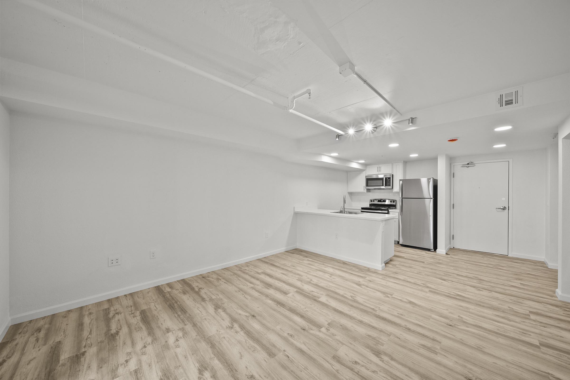 A spacious, modern studio apartment with light wood flooring. The walls are painted white, and there is a kitchenette equipped with stainless steel appliances. Recessed lighting illuminates the room, and the entrance door is visible in the background. The overall ambiance is bright and minimalistic.