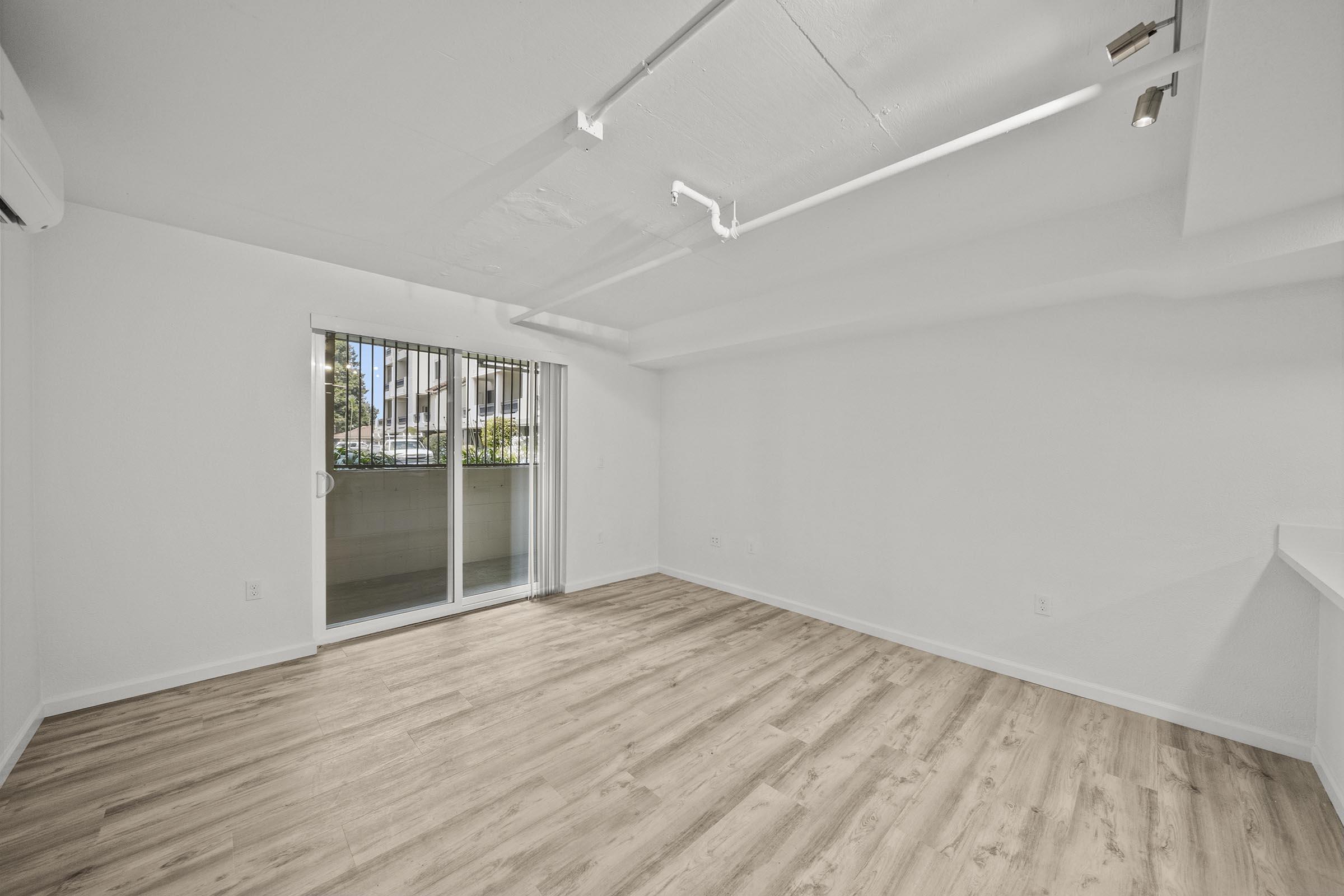 A spacious, empty room with light-colored walls and wooden flooring. There are large windows with vertical blinds, letting in natural light. The room features modern lighting fixtures and a sliding glass door leading to a balcony or outdoor area. The overall atmosphere is bright and inviting.