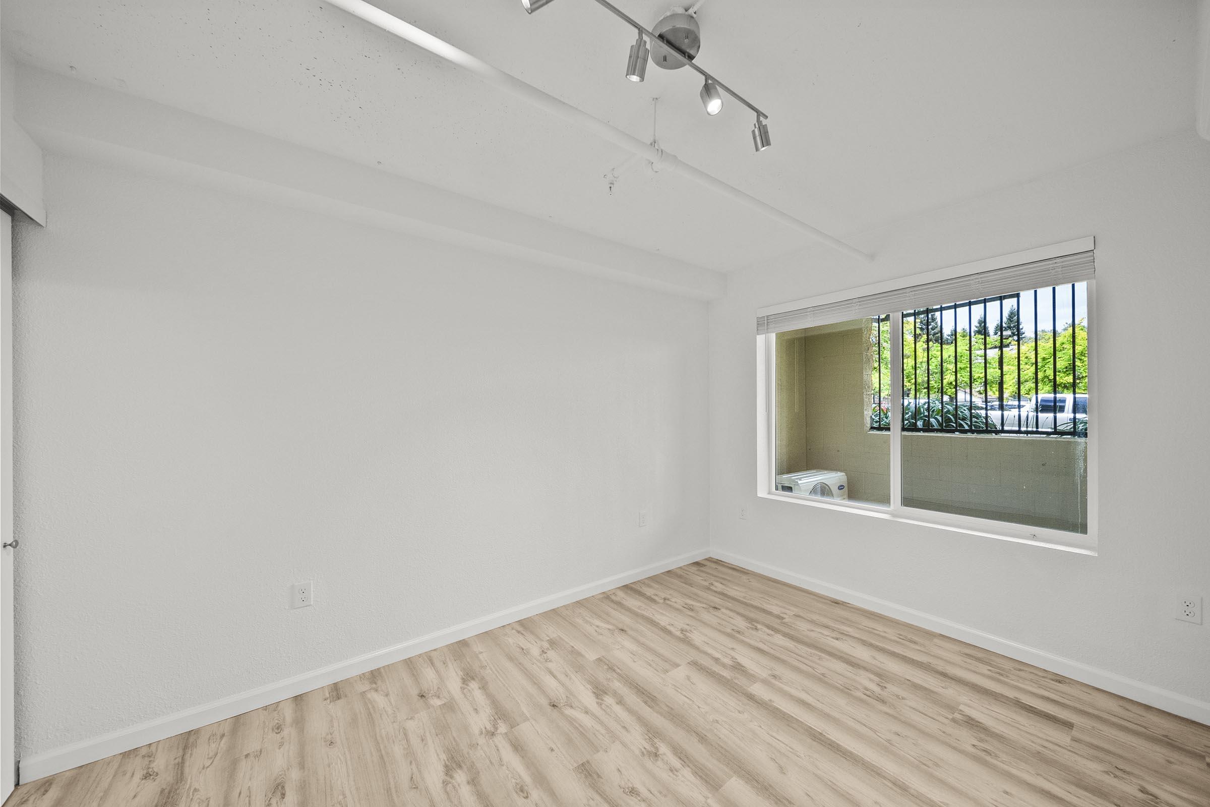 Interior of a modern, empty room featuring light-colored walls and wooden flooring. A large window allows natural light in, offering a view of green trees and an air conditioning unit outside. The ceiling is equipped with track lighting, enhancing the brightness of the space.