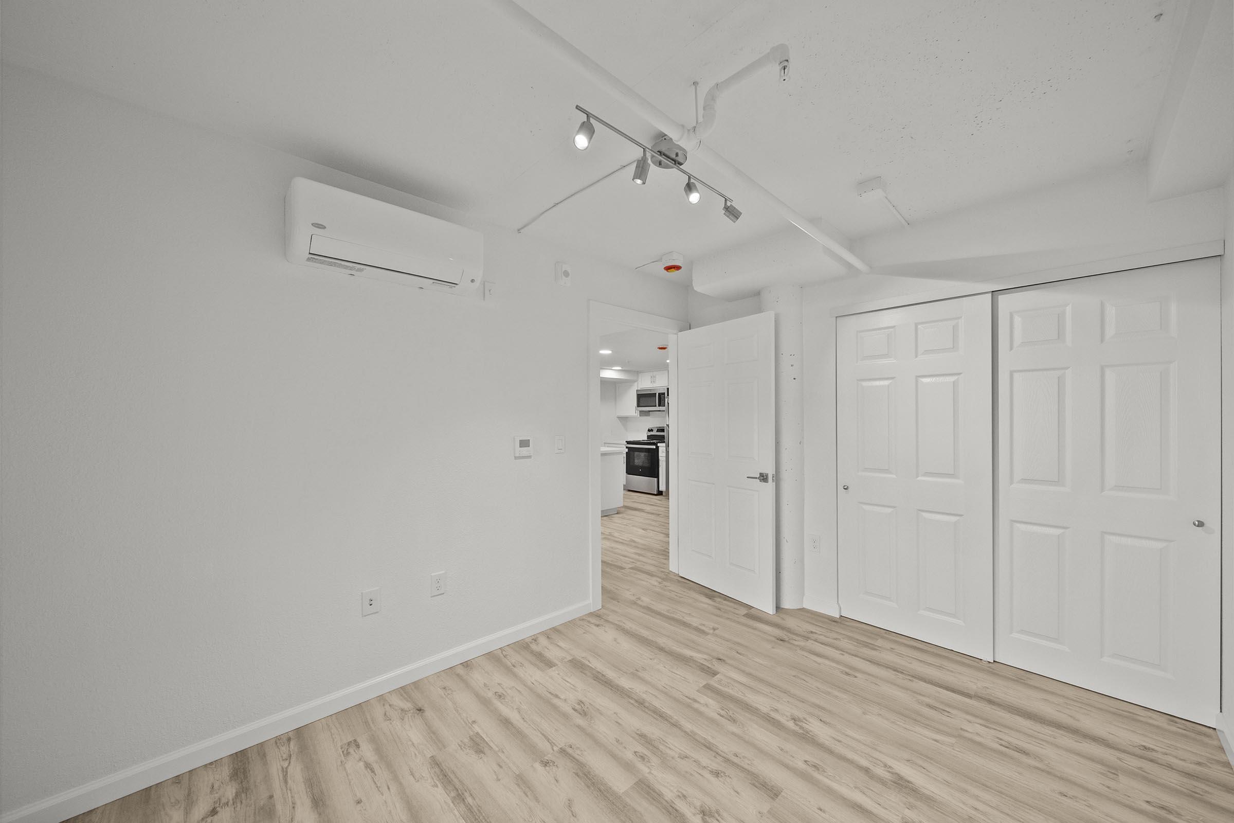 A well-lit, modern room featuring light-colored walls and wooden flooring. On the left, there is an air conditioning unit mounted on the wall, and on the right, a set of double doors leads to a closet. The room has recessed lighting and an open layout leading into a kitchen area visible in the background.
