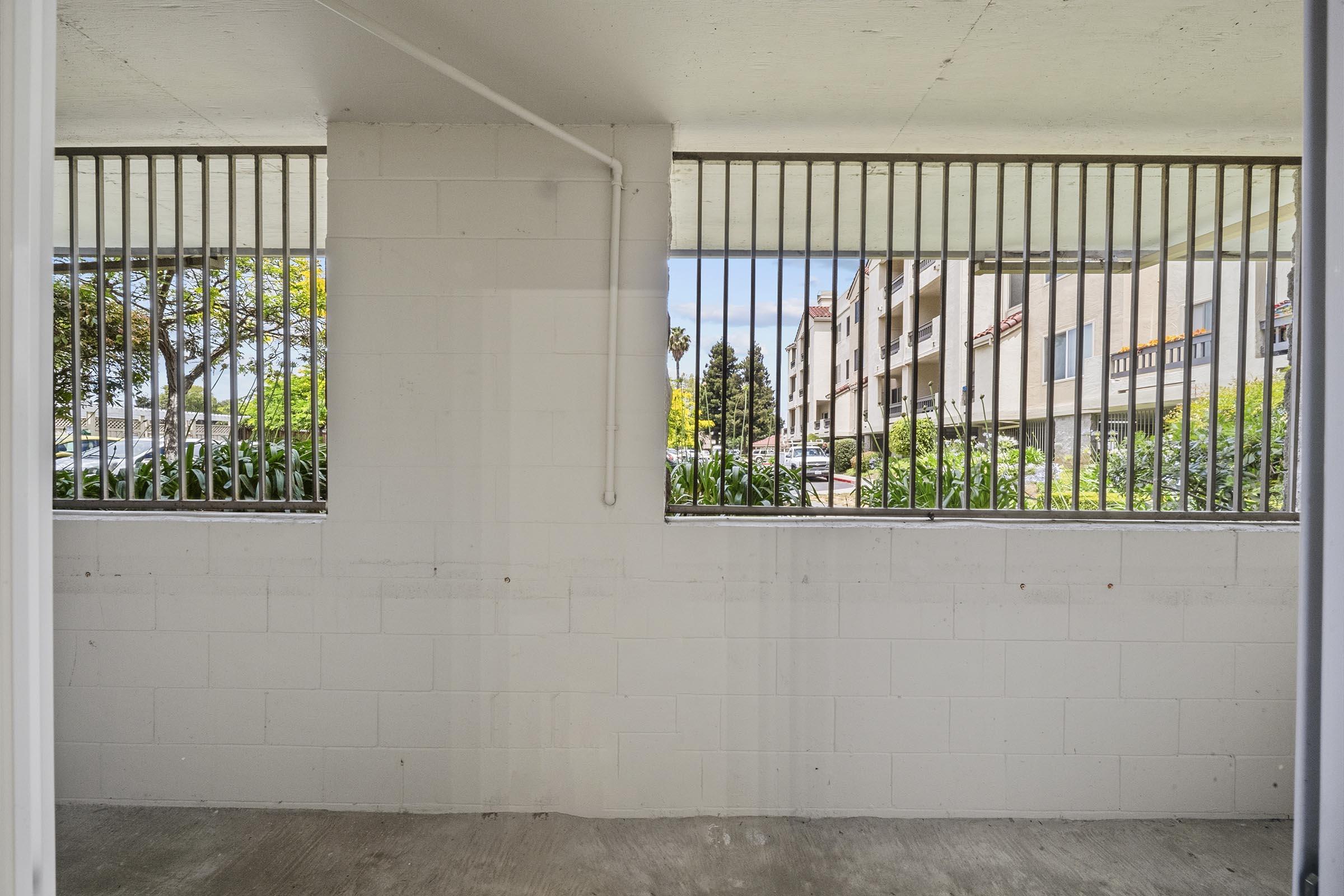View from a covered balcony showing a concrete wall with metal bars. Beyond the bars, trees and parked cars are visible, along with several buildings in the background. The sky is bright with scattered clouds.