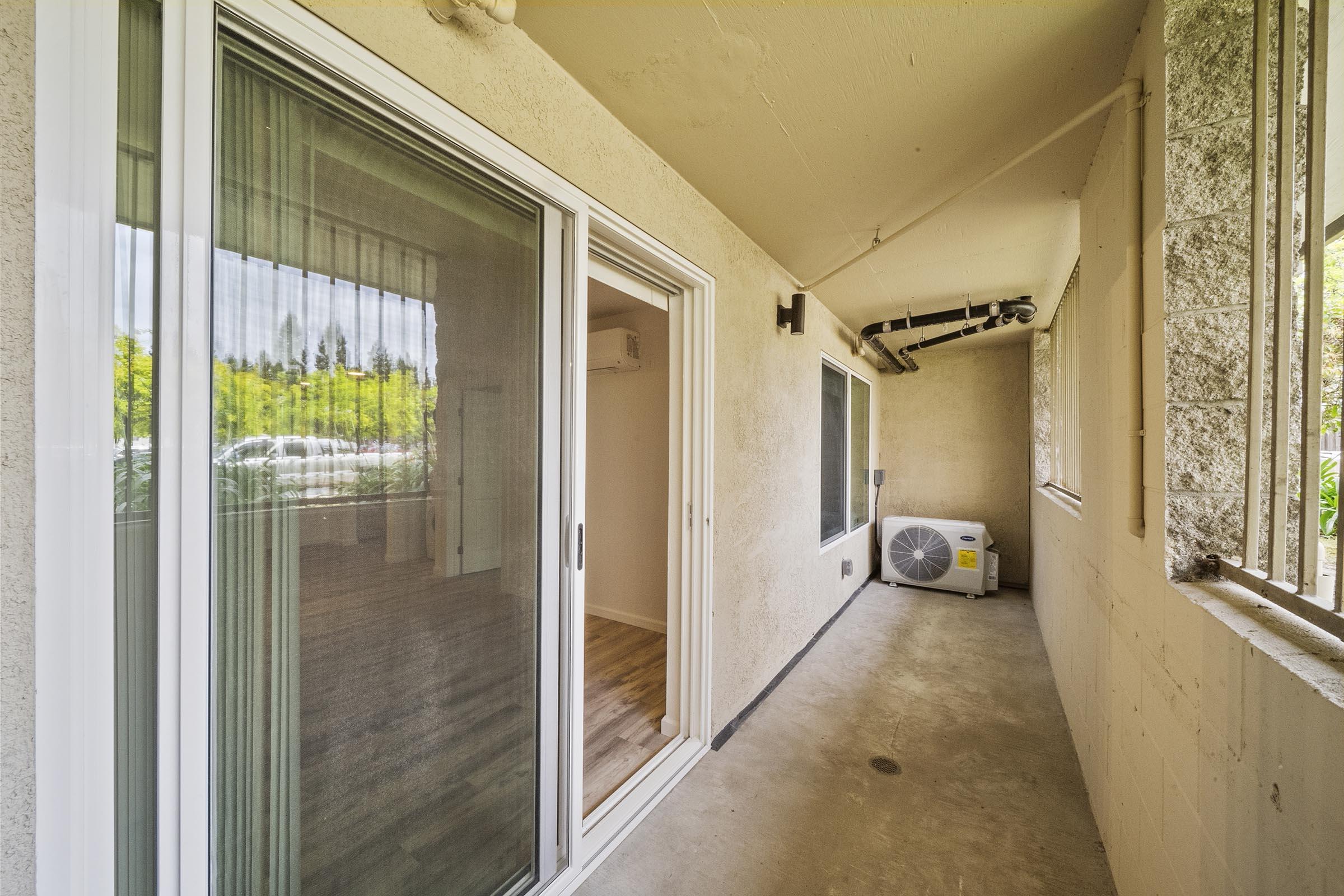 A view of a small covered balcony or porch area with a sliding glass door, concrete floor, and an air conditioning unit. The walls are beige, and there are large windows allowing natural light. The space appears to be part of an apartment or condo.