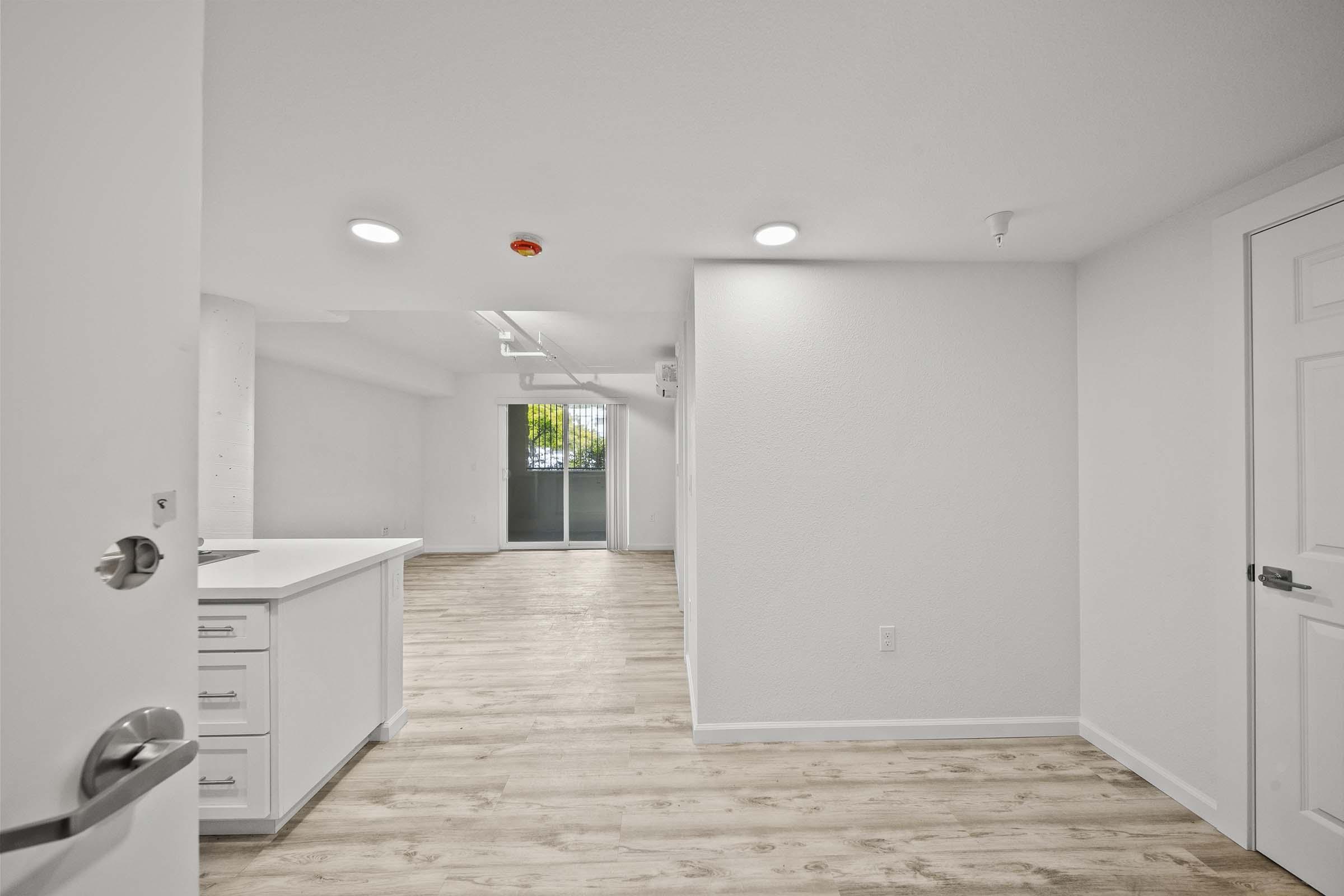 Bright, modern interior of an apartment featuring an open floor plan. The space includes a kitchen area with white cabinetry and a door leading to a patio or balcony. The walls are painted white, and flooring is light-colored wood. Ample natural light filters in through the sliding glass doors.
