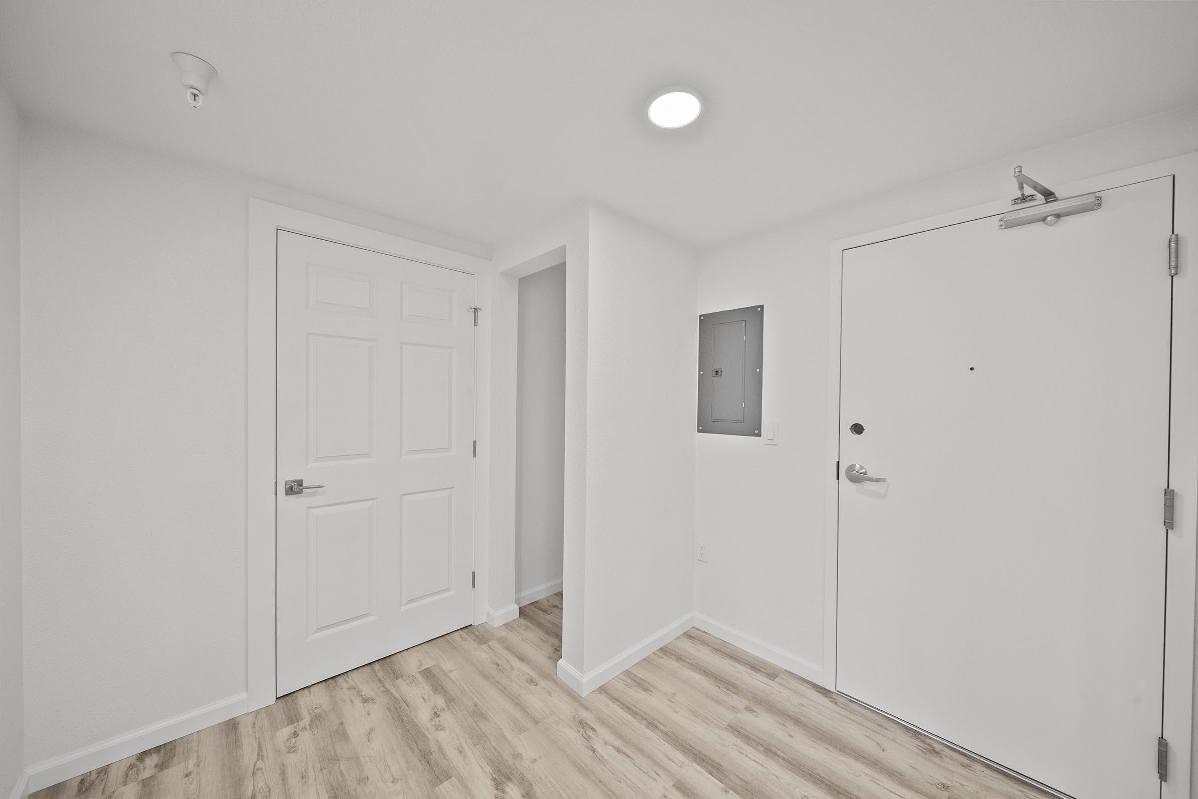 A modern, minimalist interior hallway with white walls and a light wooden floor. The space features a closed door on the left, a small closet or utility area in the center, and an entrance door on the right. A ceiling light illuminates the area, creating a bright and inviting atmosphere.