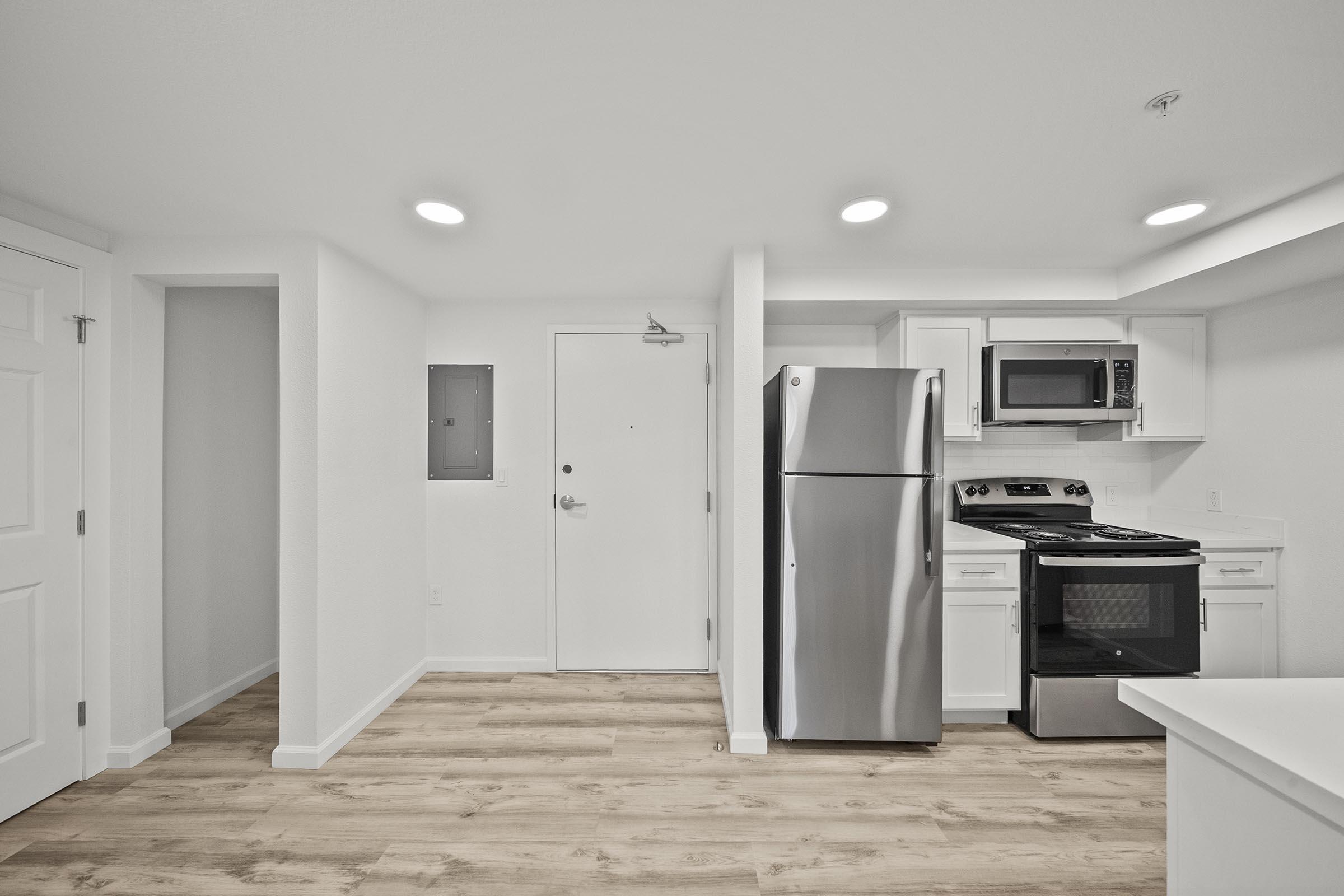 Modern kitchen and living area featuring a stainless steel refrigerator, microwave, and stove. The space has light-colored cabinetry and a neutral color scheme with a door leading outside. Natural wood flooring enhances the contemporary look.