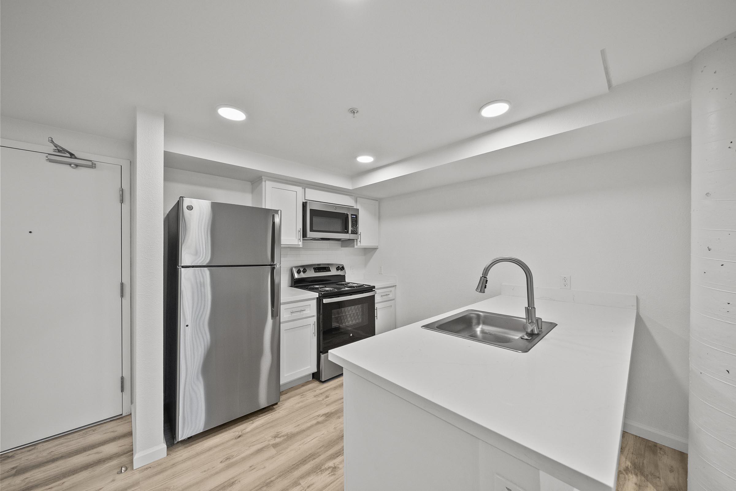 Modern kitchen featuring a stainless steel refrigerator, microwave, and stove, with white cabinets and a sleek countertop. Neutral-colored walls and flooring enhance the spacious feel, complemented by bright overhead lighting.